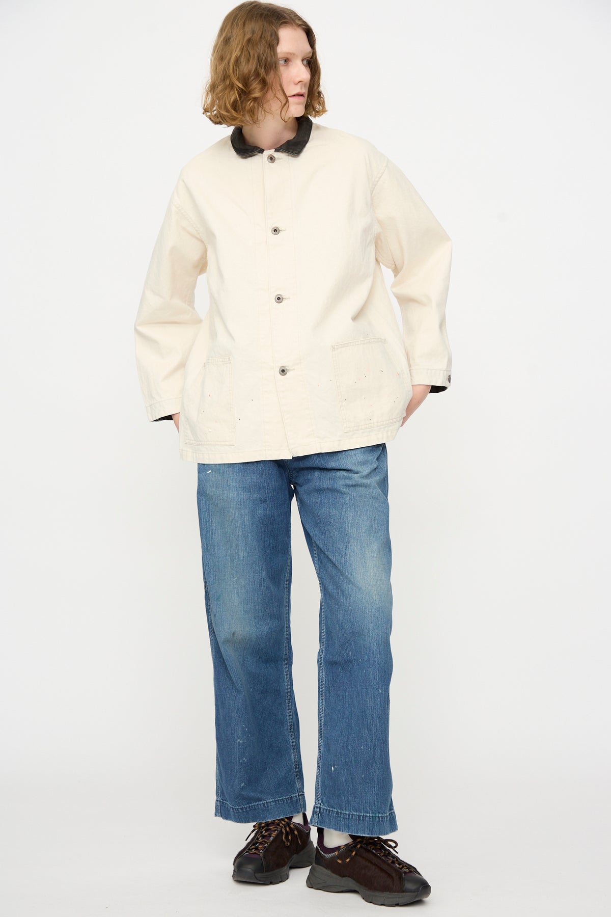 A person stands against a plain background, wearing the Chimala Twill Engineer Jacket in Ivory Splatter made in Japan, paired with blue jeans and dark sneakers; their hands are in their pockets and their head is turned slightly to the side.