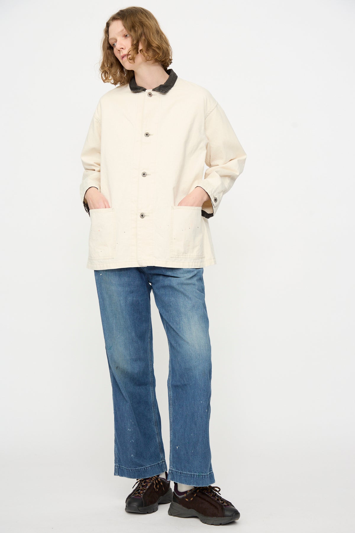 A person stands against a white background, wearing the Chimala Twill Engineer Jacket in Ivory Splatter, paired with blue jeans and black shoes, with hands in the jacket’s pockets.