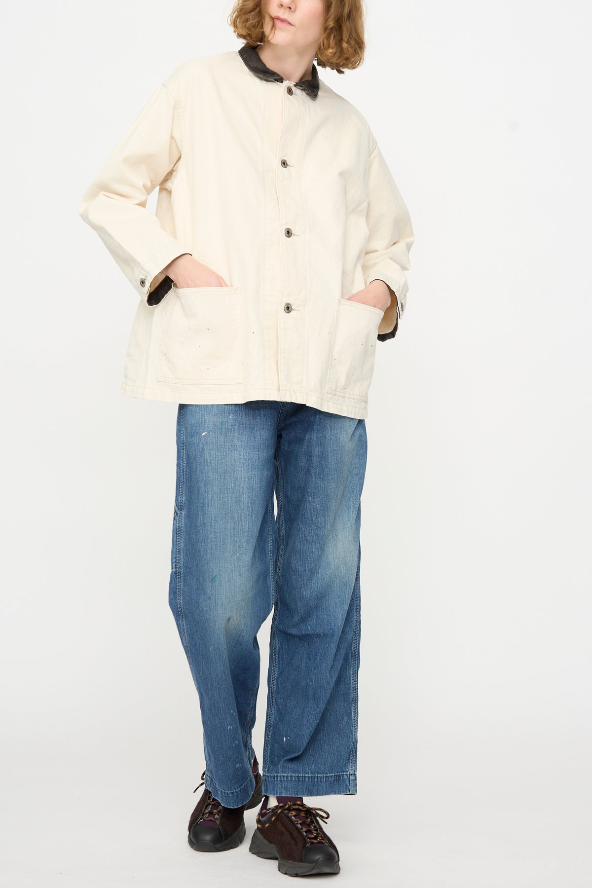 A person wearing the Chimala Twill Engineer Jacket in Ivory Splatter, paired with blue wide-leg jeans and black shoes, stands with hands in jacket pockets against a plain white background. Expertly tailored in Japan, the jacket showcases refined craftsmanship and elevates the effortlessly stylish look.