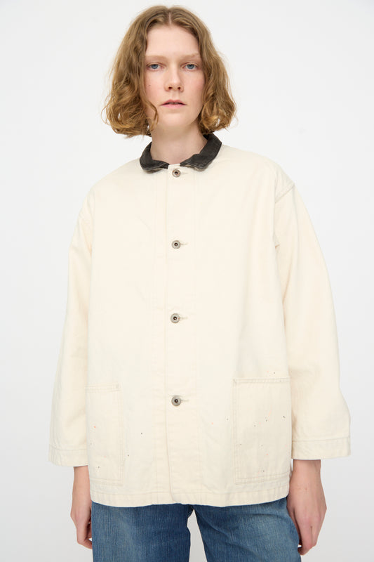 A person with shoulder-length blonde hair is seen wearing the Chimala Twill Engineer Jacket in Ivory Splatter, a Japanese-made jacket featuring front pockets and a dark collar, paired with blue jeans, standing against a plain light background.