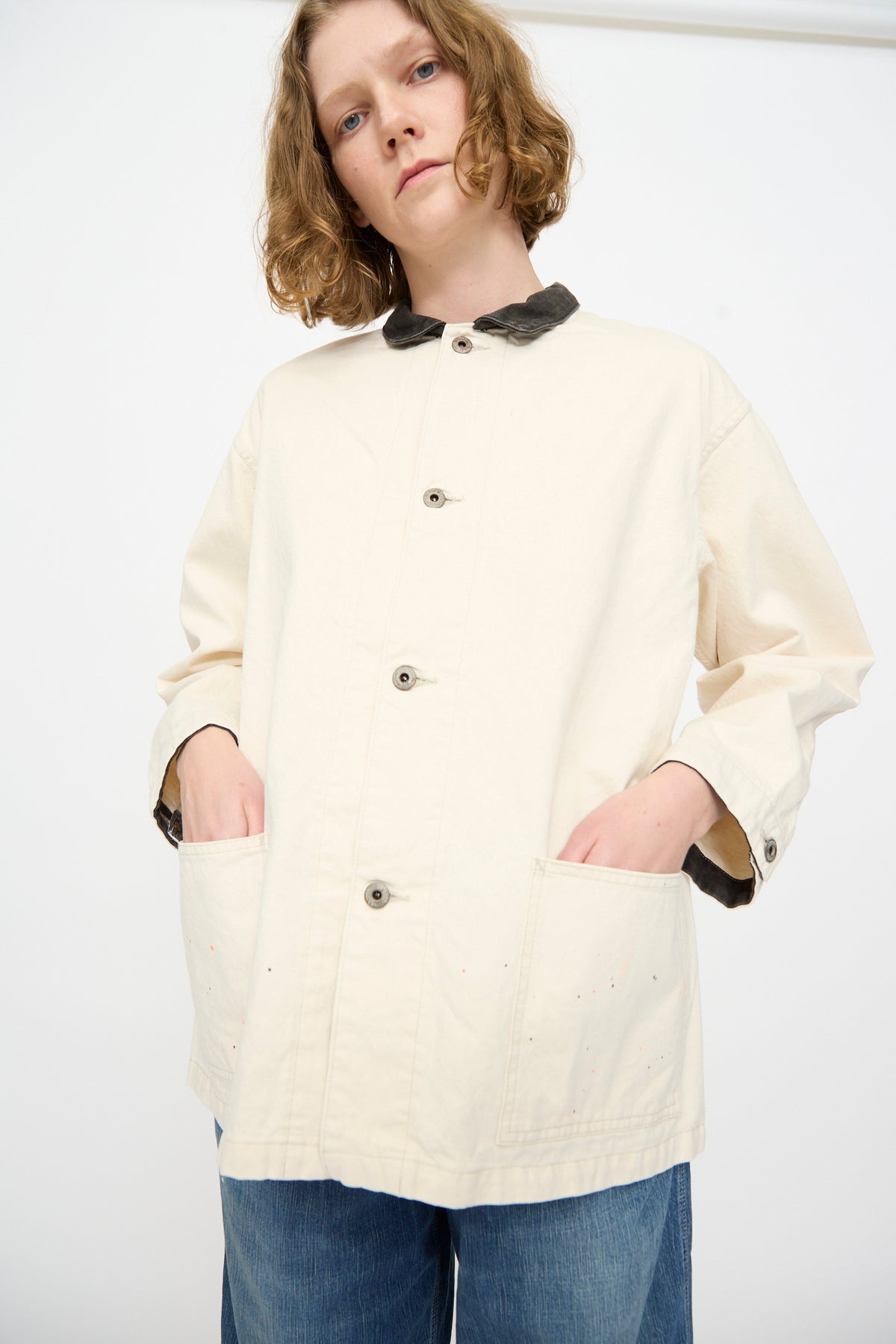 A person wears the Chimala Twill Engineer Jacket in Ivory Splatter, featuring large pockets and a contrasting dark collar, crafted in Japan. They stand with hands in pockets, looking slightly down at the camera.