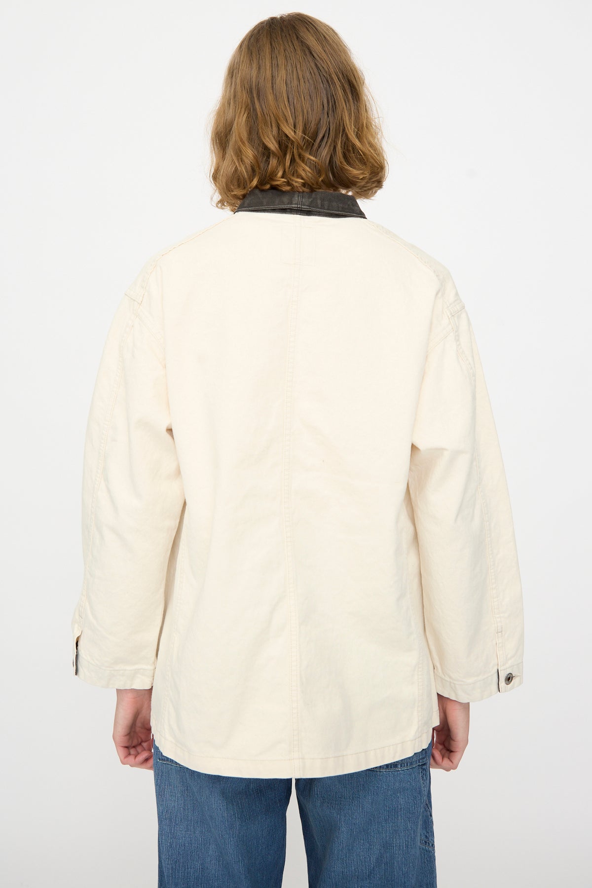 A person with shoulder-length hair is shown from the back against a plain white background, wearing Chimala’s Twill Engineer Jacket in Ivory Splatter, featuring a dark collar and paired with blue jeans. This jacket is expertly made in Japan.