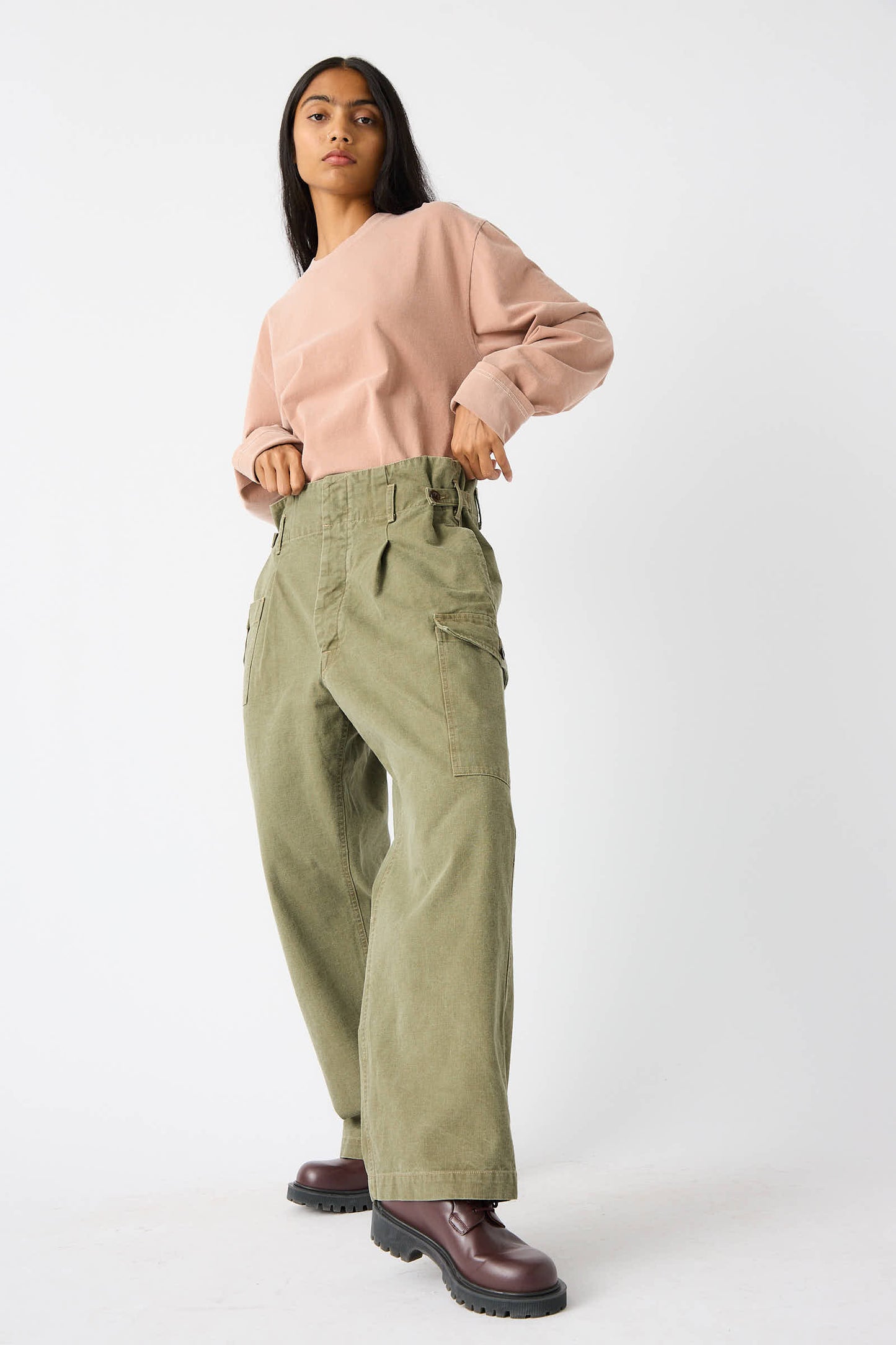 A person stands against a plain white background, wearing a long-sleeve pink top and relaxed fit, mid-rise Unisex Belgian Waist Gathering Cargo Pant in Olive from Chimala, crafted from sulfur-dyed canvas. Dark brown boots complete the look as they adjust their top.