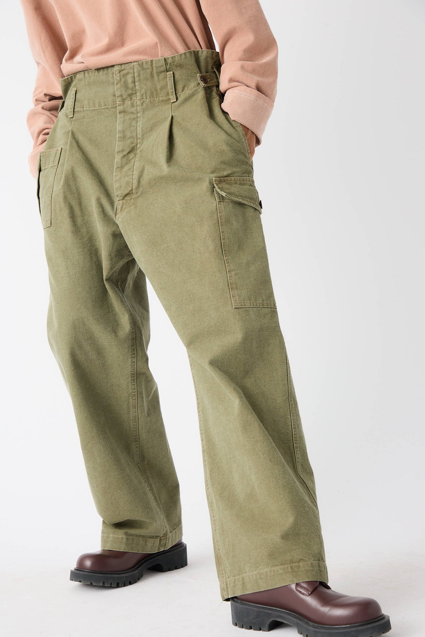 Person wearing Chimala's Unisex Belgian Waist Gathering Cargo Pant in Olive with a relaxed fit and hands in pockets, paired with a beige top and brown boots, standing against a plain white background.