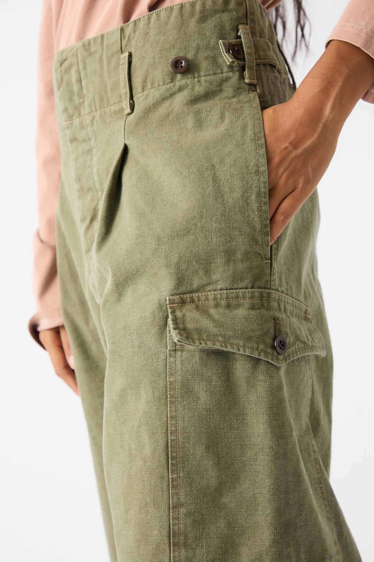 A person wearing Chimala's Unisex Belgian Waist Gathering Cargo Pant in Olive, made from sulfur-dyed canvas, with one hand in the pocket. The relaxed-fit, high-waisted pants feature belt loops, pleats, and side pockets with buttoned flaps. The person is also wearing a long-sleeve top.