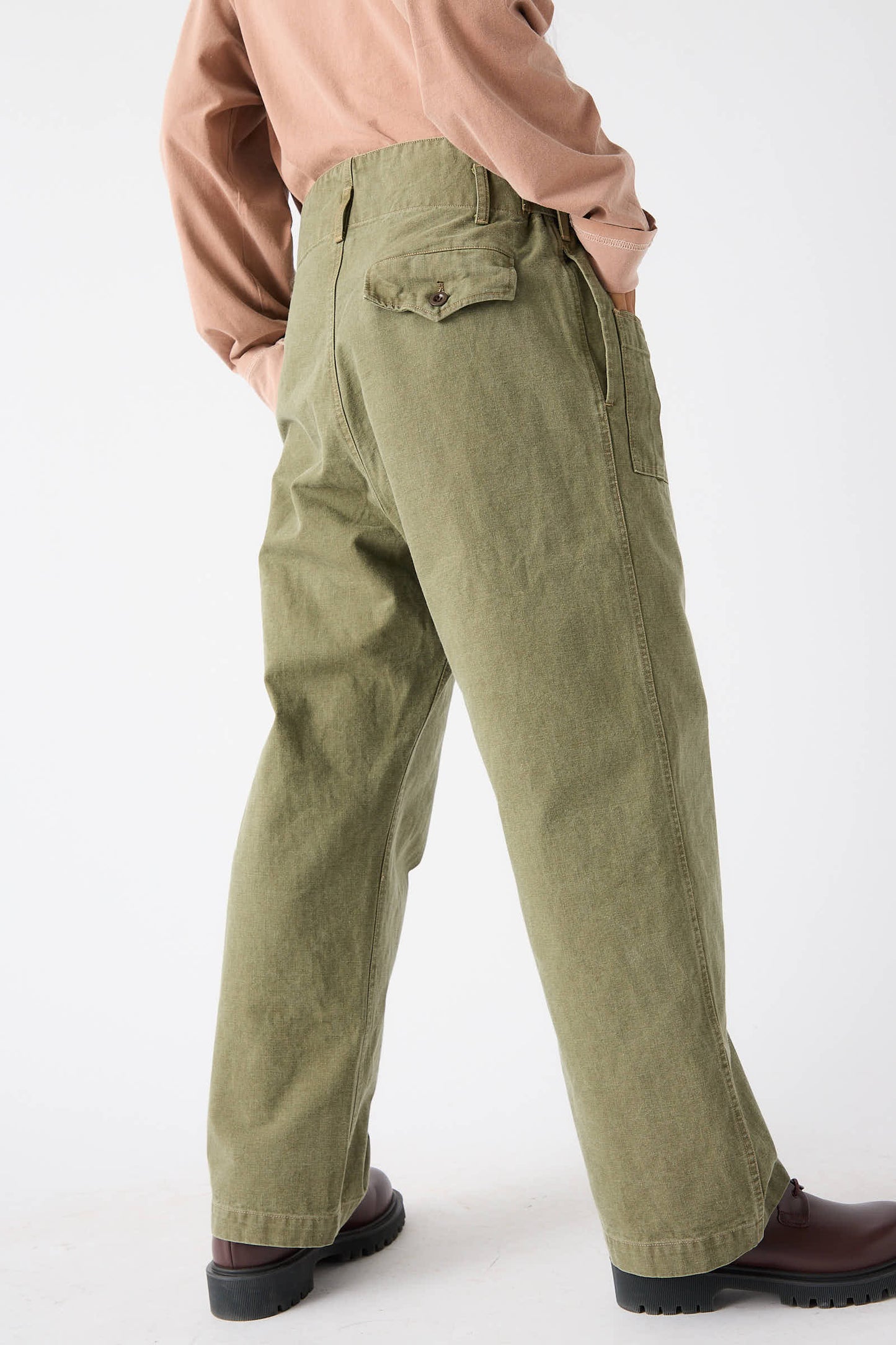 A person wearing Chimala's Unisex Belgian Waist Gathering Cargo Pant in Olive, along with a light brown shirt and dark brown shoes, is standing with their back facing the camera.