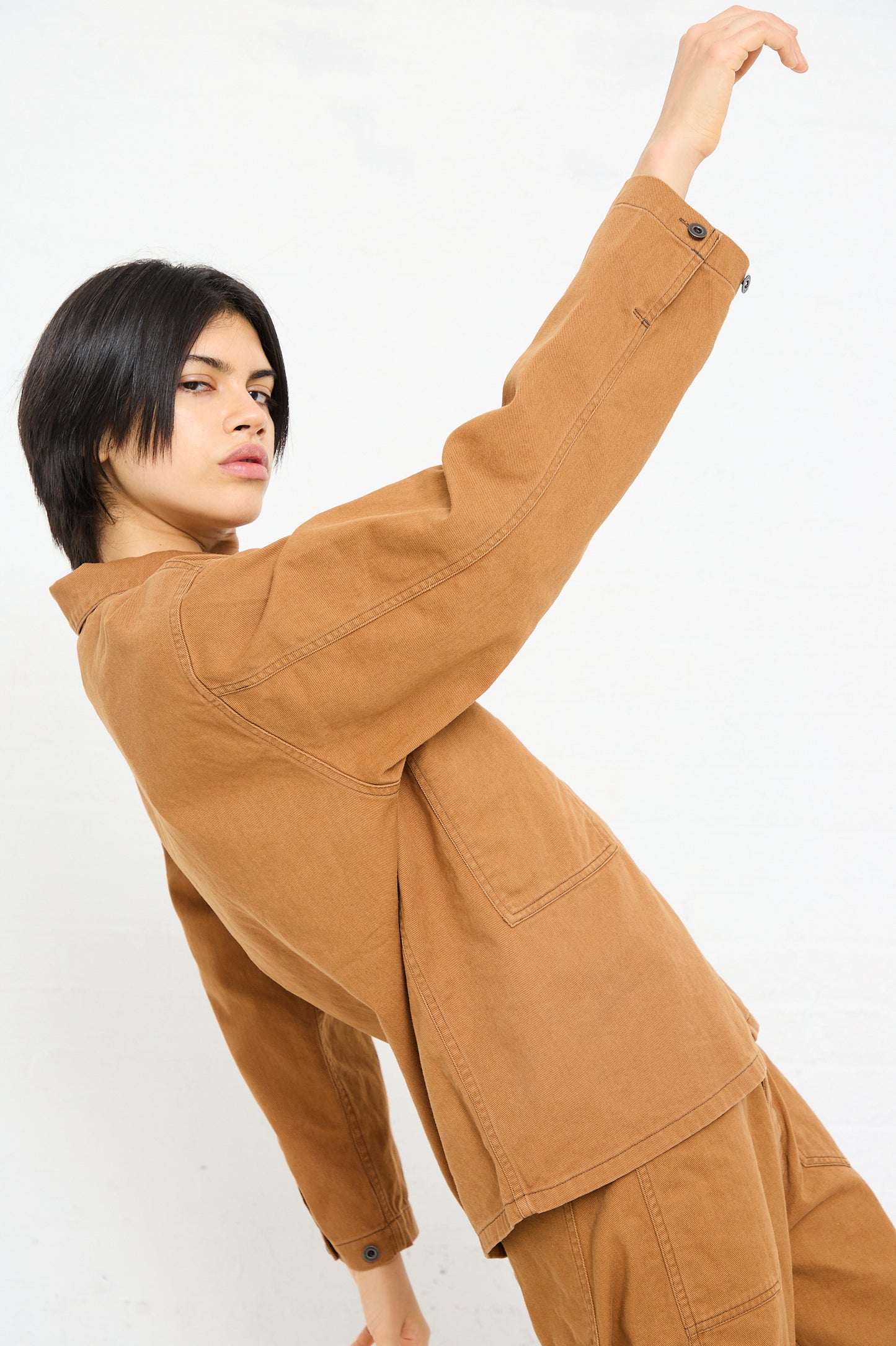 A person in a Chimala Unisex Classic Drill US Army Work Jacket in Camel posing with one arm stretched upward against a white background.