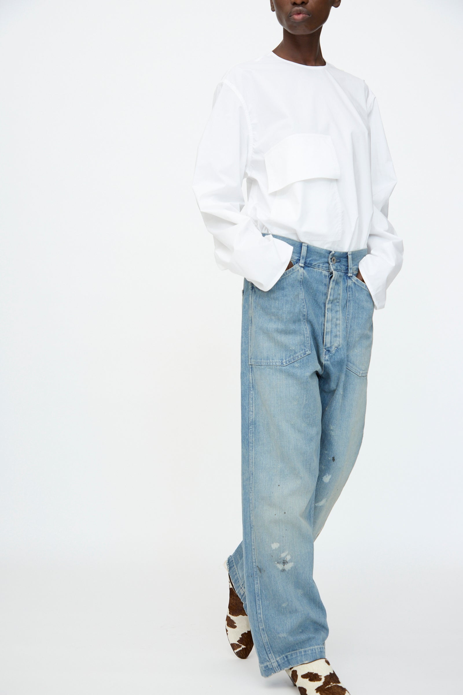 A person is wearing a white long-sleeve shirt, Chimala Unisex US Army Style Denim Trouser in Light Vintage, and brown-and-white patterned shoes, standing against a plain background.