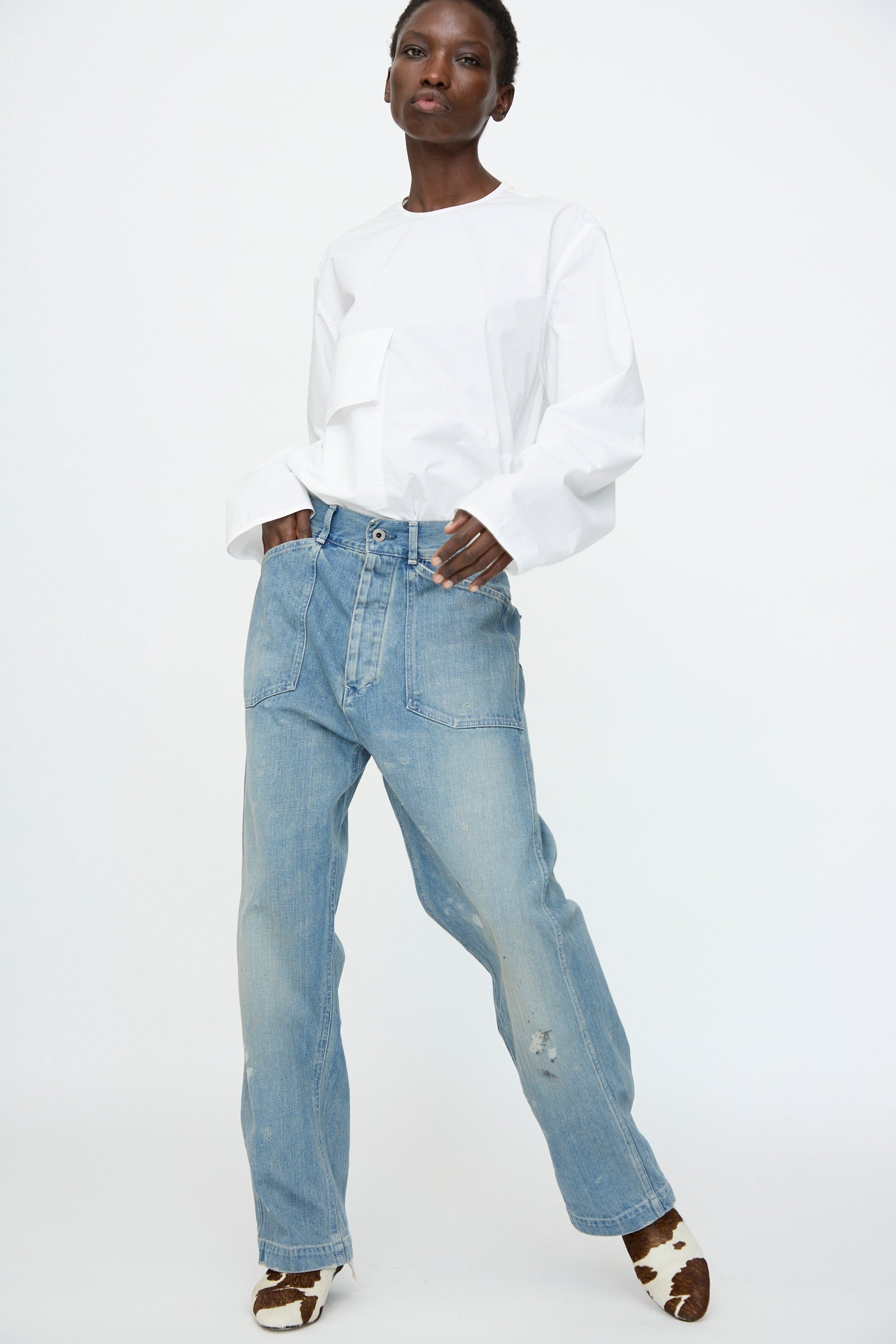 A person wearing a white long-sleeve shirt, Chimala Unisex US Army Style Denim Trouser in Light Vintage, and brown-and-white patterned shoes stands against a plain white background.