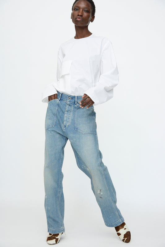A person wearing a white long-sleeve shirt, Chimala Unisex US Army Style Denim Trouser in Light Vintage, and brown-and-white patterned shoes stands against a plain white background.