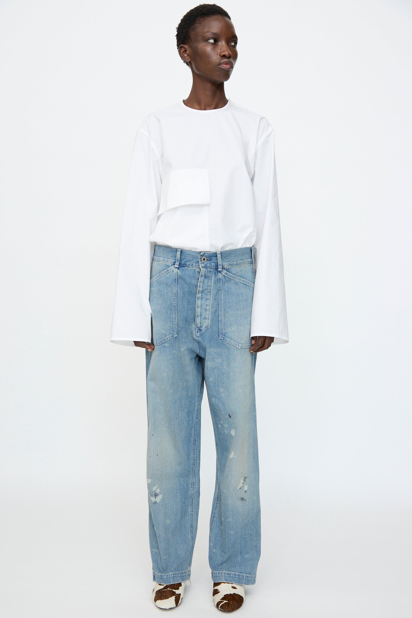 Person standing against a plain background, wearing a long-sleeved white top with a front pocket, Chimala Unisex US Army Style Denim Trouser in Light Vintage, and patterned heeled shoes.