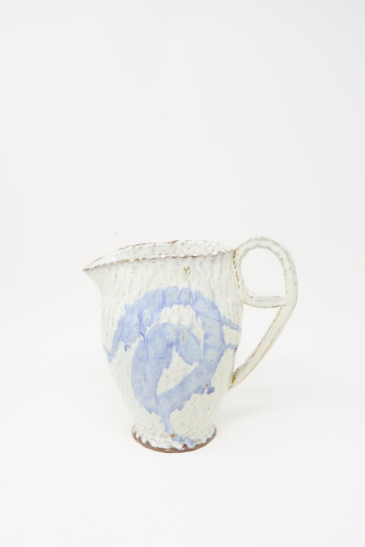 The Ceramic Pitcher in Blue by Claire Resnick Ceramics is a handmade ceramic piece featuring a textured cream surface with a light blue swirled pattern, a large handle, and a small spout, displayed on a plain white background.