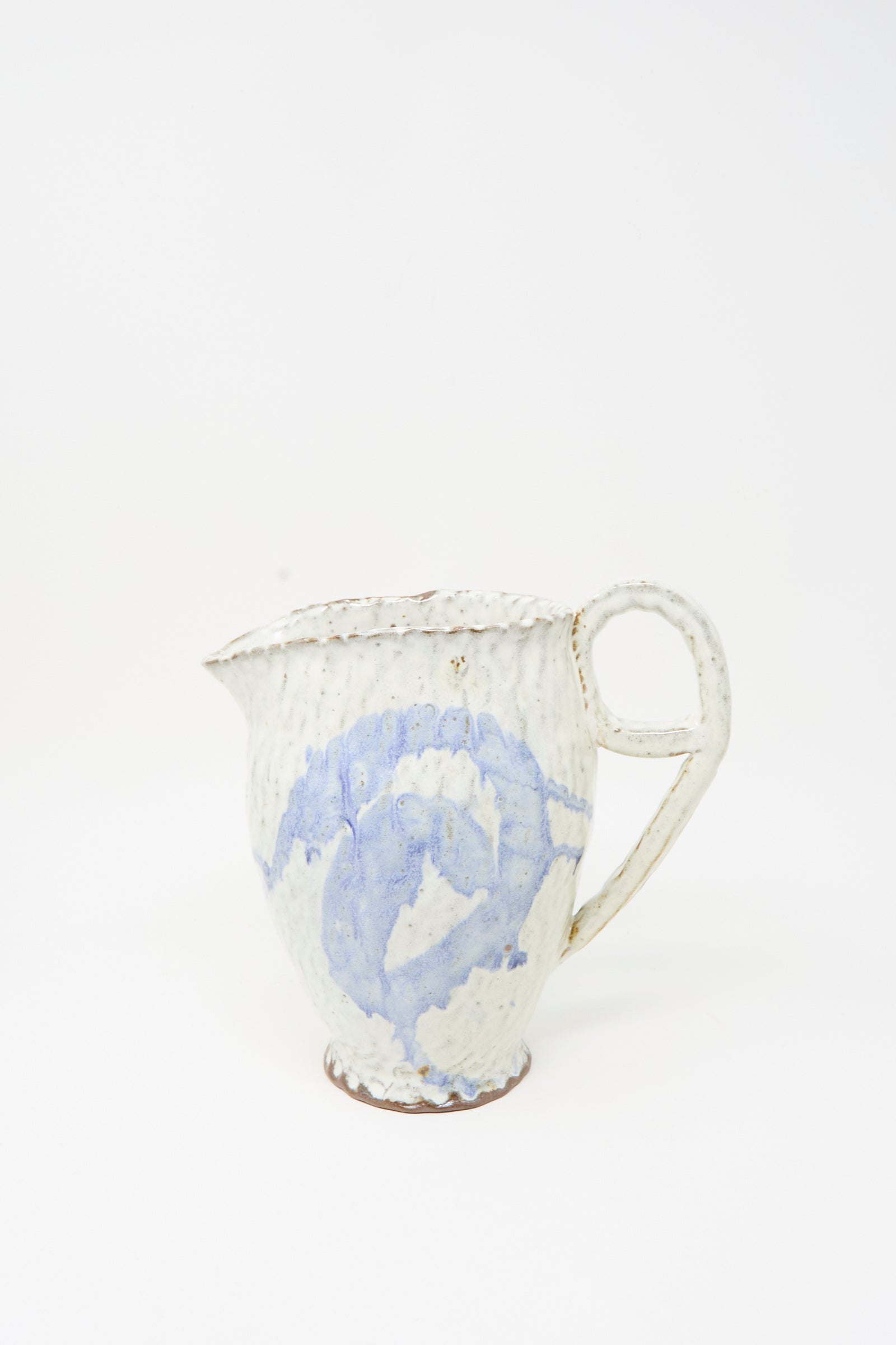 The Ceramic Pitcher in Blue by Claire Resnick Ceramics is a handmade ceramic piece featuring a textured cream surface with a light blue swirled pattern, a large handle, and a small spout, displayed on a plain white background.