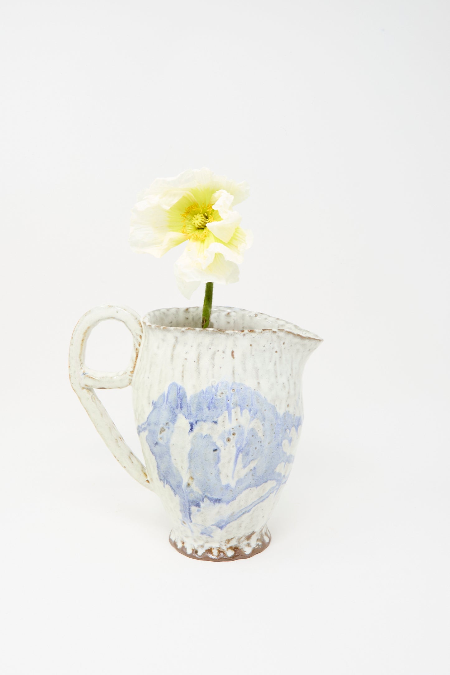 A solitary pale yellow poppy is arranged in the Claire Resnick Ceramics “Ceramic Pitcher in Blue,” a handmade white and blue vessel featuring a delicate blue glaze, photographed against a plain white background.