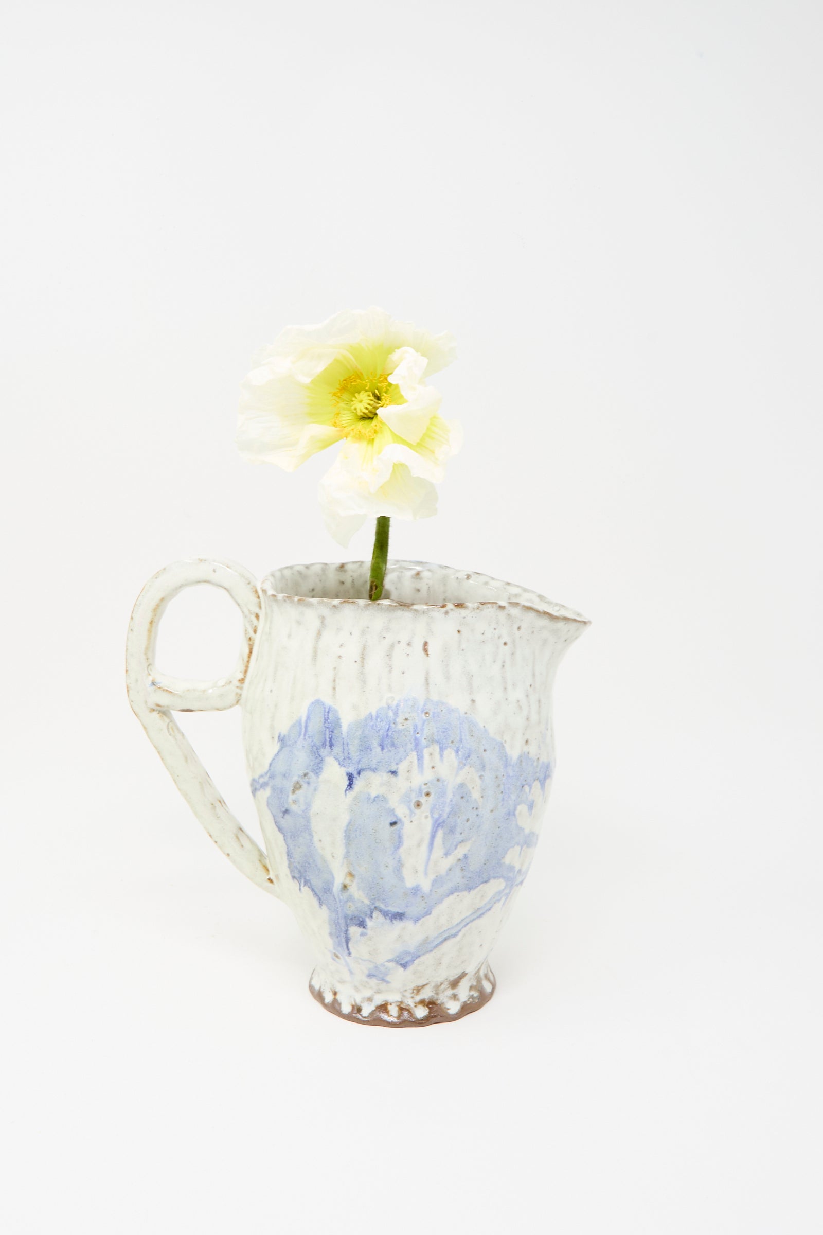 A solitary pale yellow poppy is arranged in the Claire Resnick Ceramics “Ceramic Pitcher in Blue,” a handmade white and blue vessel featuring a delicate blue glaze, photographed against a plain white background.