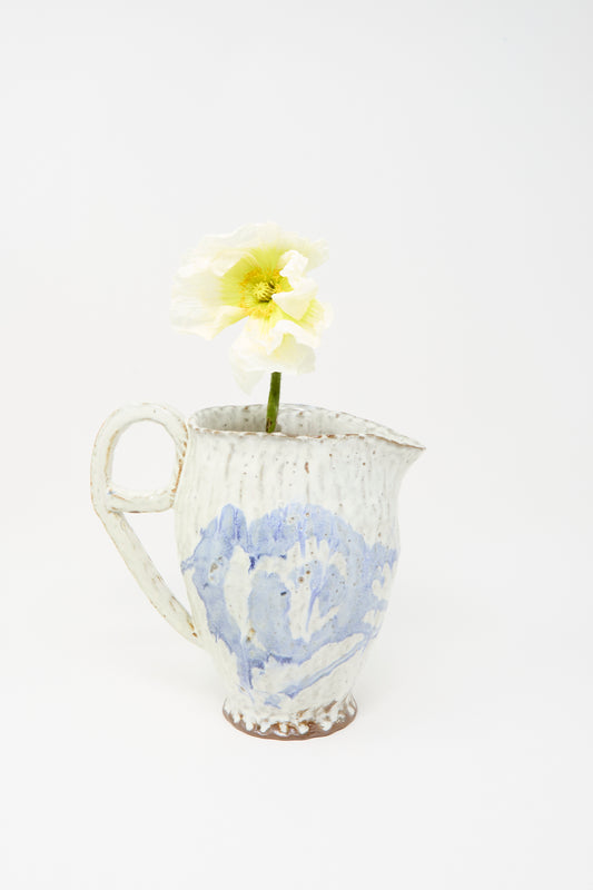 A solitary pale yellow poppy is arranged in the Claire Resnick Ceramics “Ceramic Pitcher in Blue,” a handmade white and blue vessel featuring a delicate blue glaze, photographed against a plain white background.