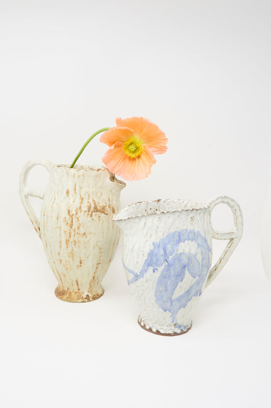 Two Claire Resnick Ceramics pitchers in tan, hand-built in the USA, are placed side by side; one features cream coloring and the other has blue accents. The cream pitcher displays a single peach poppy flower with a green stem. Plain white background.