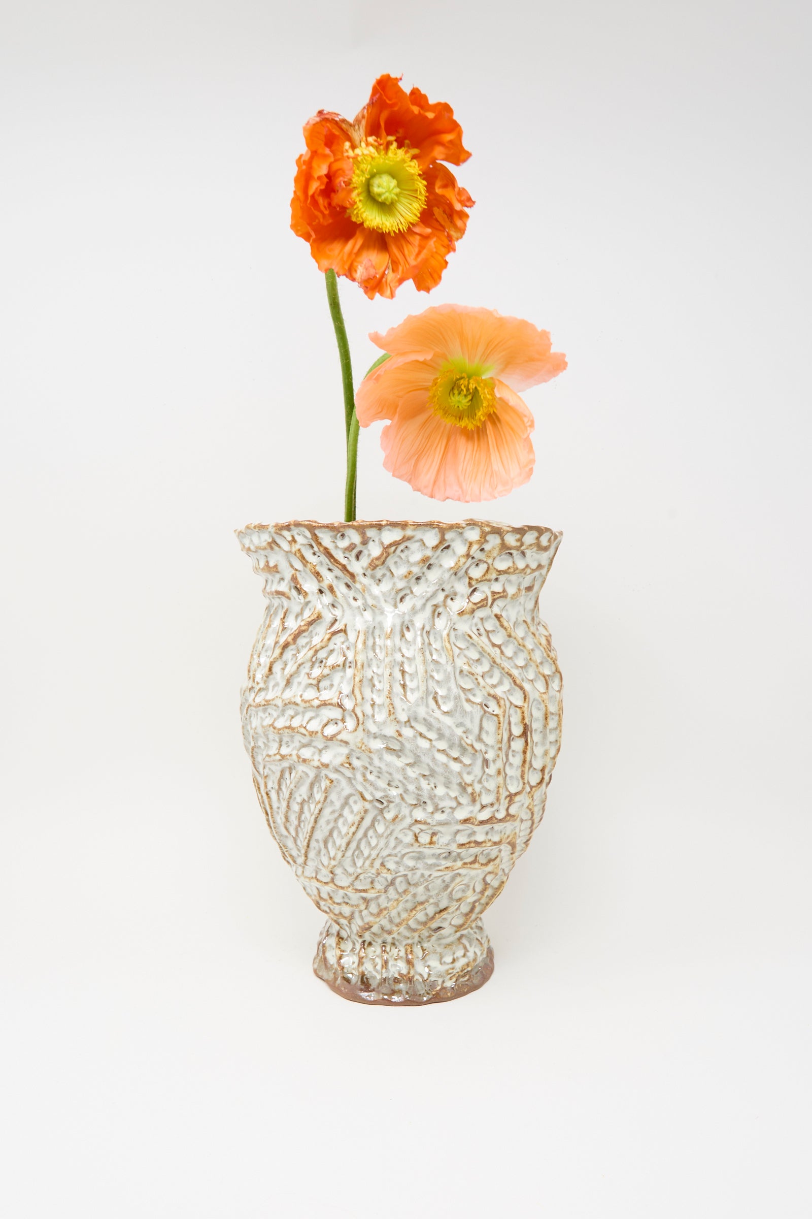 The Ridge Vase in Brown by Claire Resnick Ceramics, a textured hand-built ceramic piece, holds two poppy flowers—one orange and one pale peach—set against a plain white background.