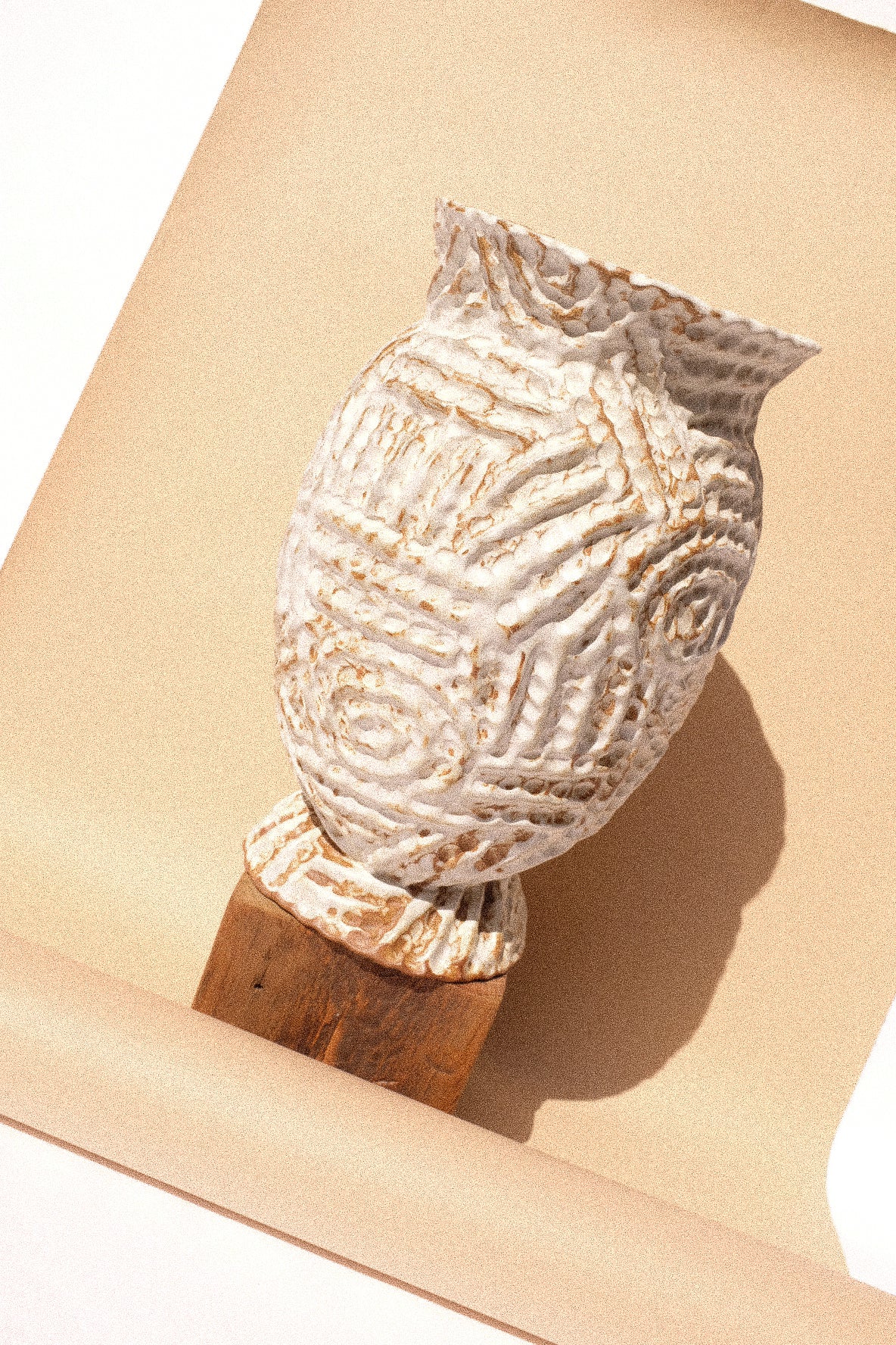 A textured ceramic owl figurine in cream and brown tones, mounted on a wooden base, set against a warm beige background with a sharp shadow.