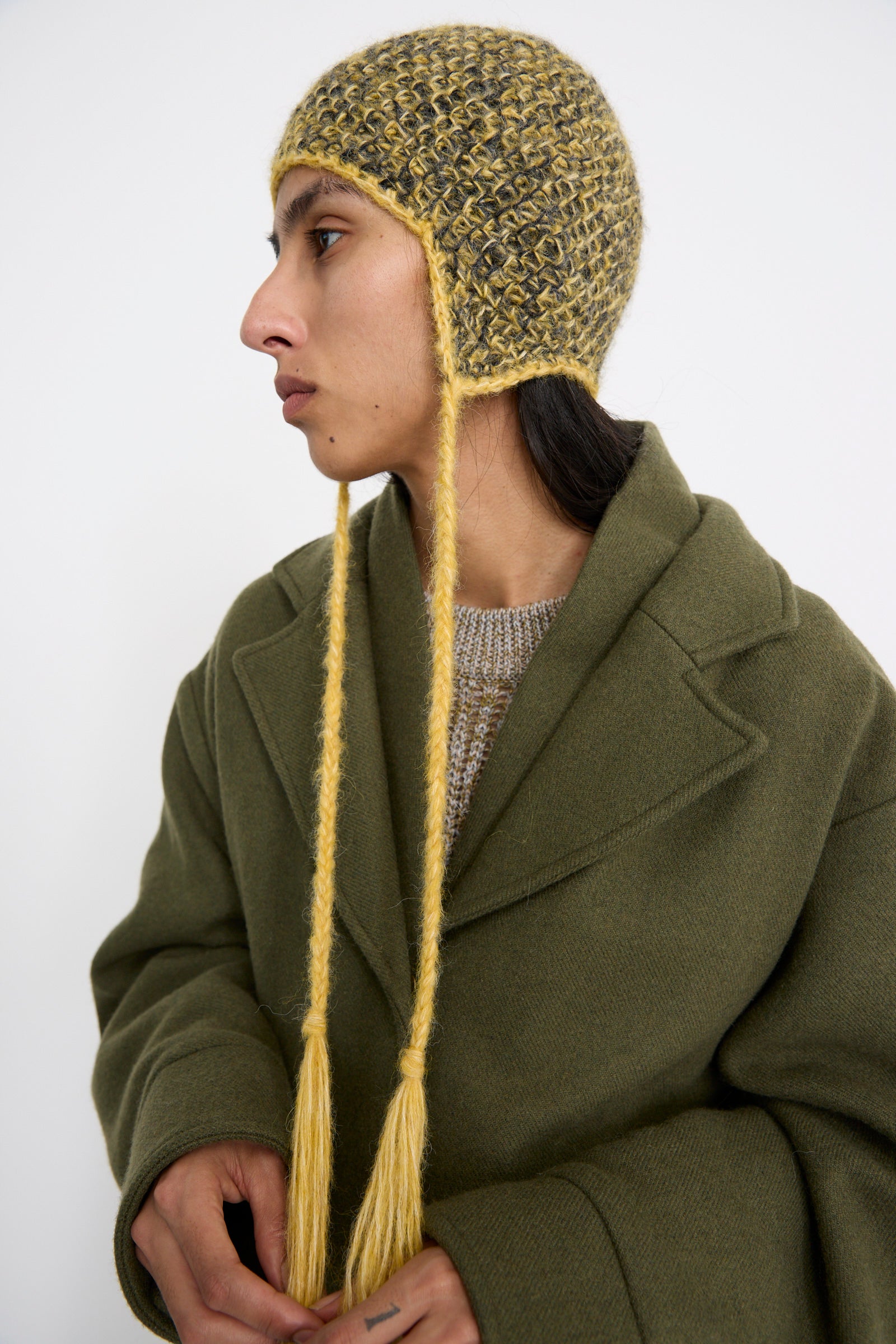 A person wearing a green coat and an Earflap Beanie in Yellow Charcoal Melange by Clyde, looking to the side against a white background. 