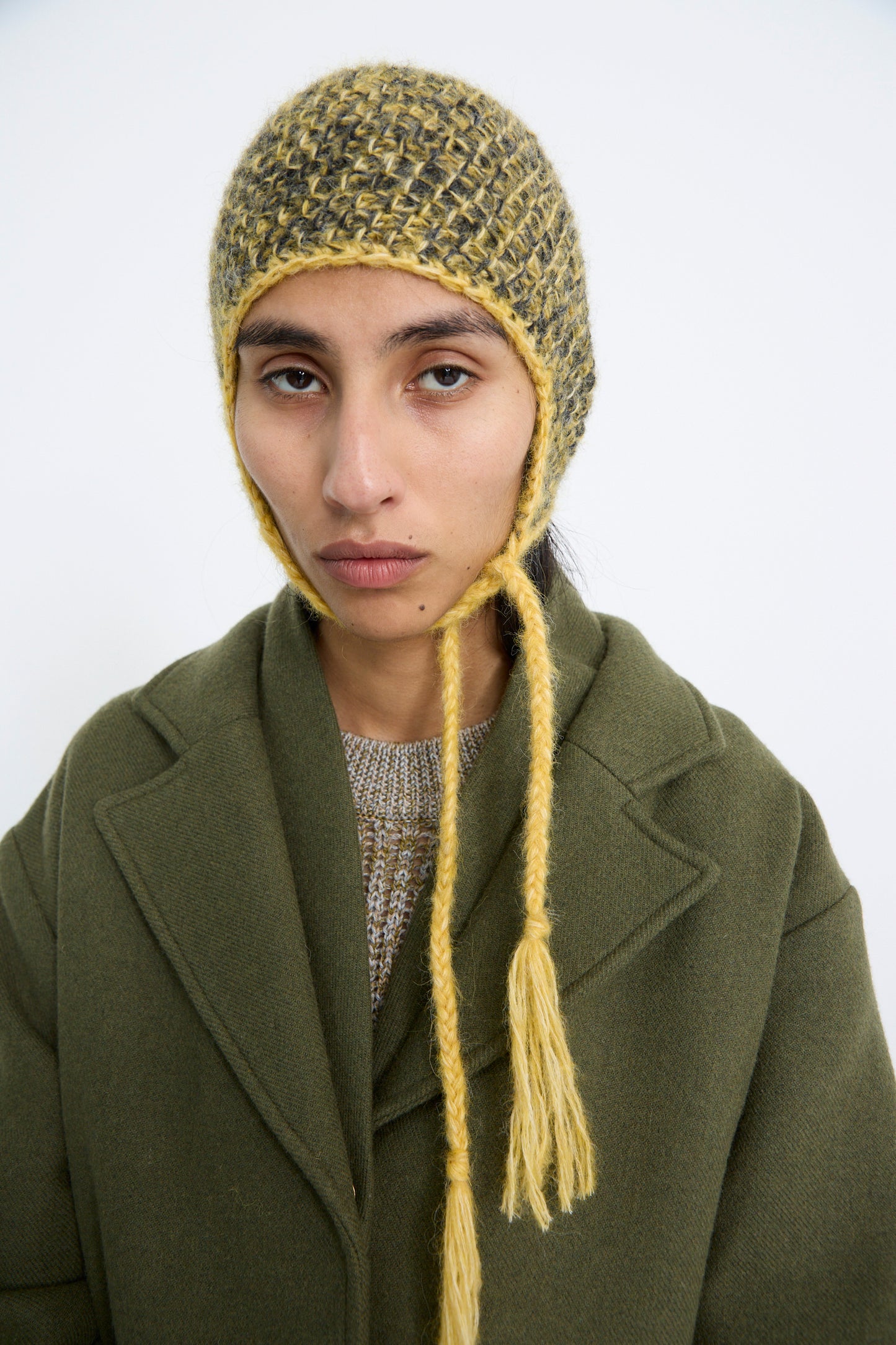 A person dressed in an olive green coat stands against a plain background, wearing Clyde's Earflap Beanie in Yellow Charcoal Melange.