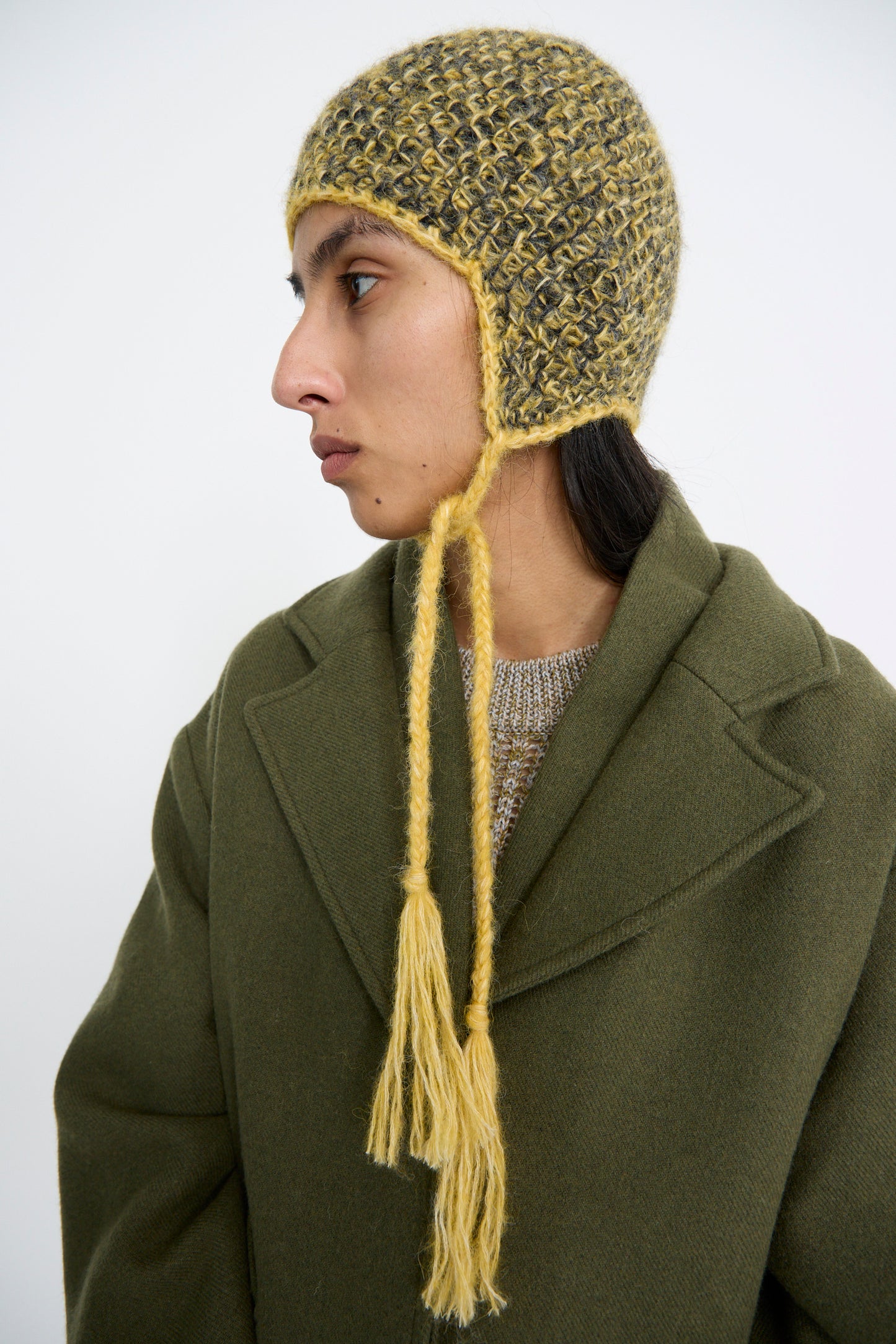 A person wearing a green coat and a Clyde Earflap Beanie in Yellow Charcoal Melange looks to the side against a plain background.