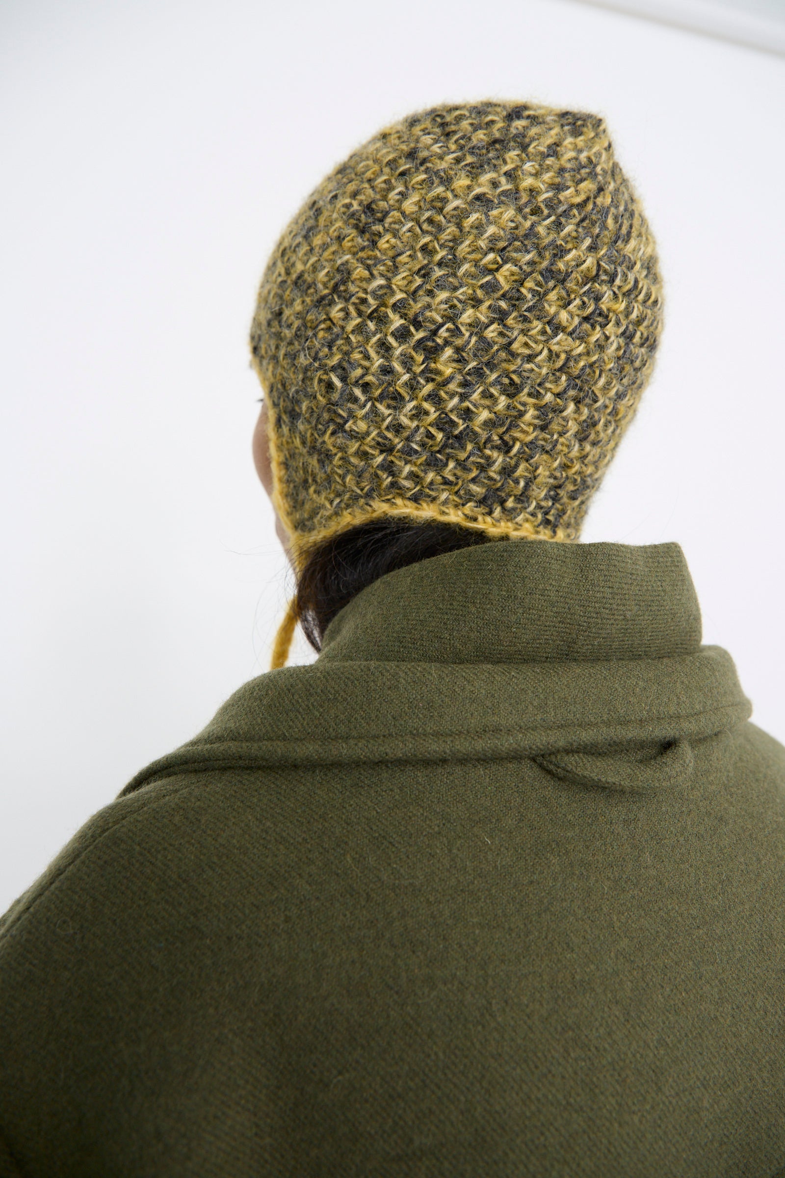 A person in a green coat, viewed from behind, is wearing a Clyde Earflap Beanie in Yellow Charcoal Melange.