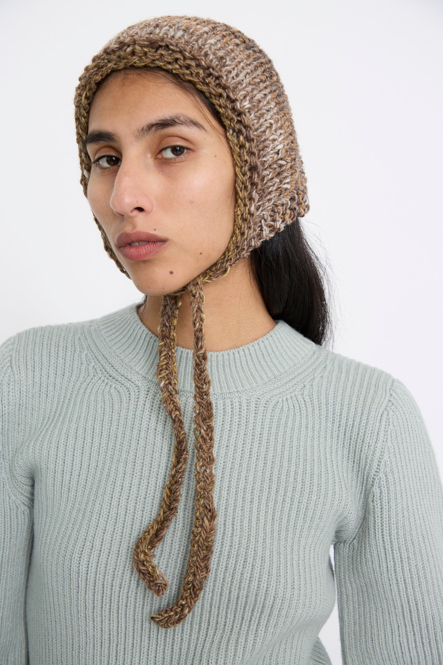 A person is sporting the Clyde Knit Wool Space Bonnet in Venus Brown, paired with a light gray sweater, as they gaze at the camera against a neutral background.