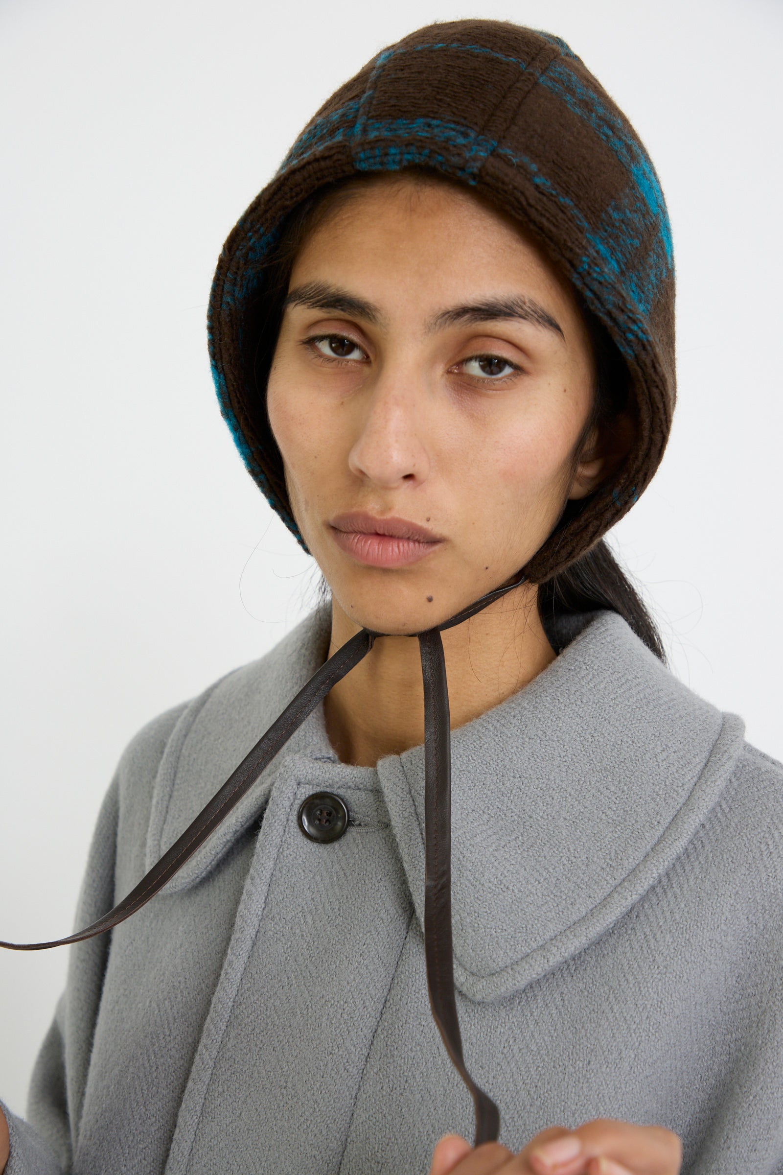 A person wearing the Clyde Volo Hat in Brown Electric Plaid and a gray coat, holding the hat strings, against a white background.