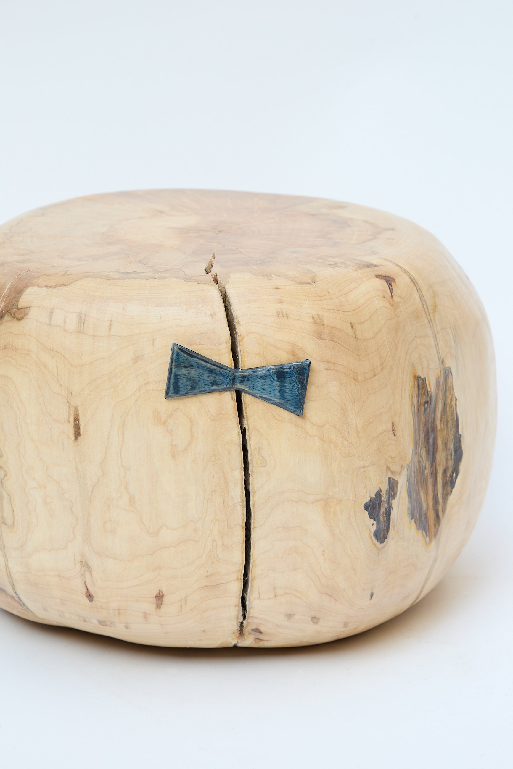 A Cody Brgant Fort Greene Maple Pouf, a round, light wooden object sculpted from a natural maple tree, featuring a natural crack held together by a blue bow tie-shaped piece on its side, revealing intricate wood grain and texture. Handmade in Brooklyn for an exquisite touch.