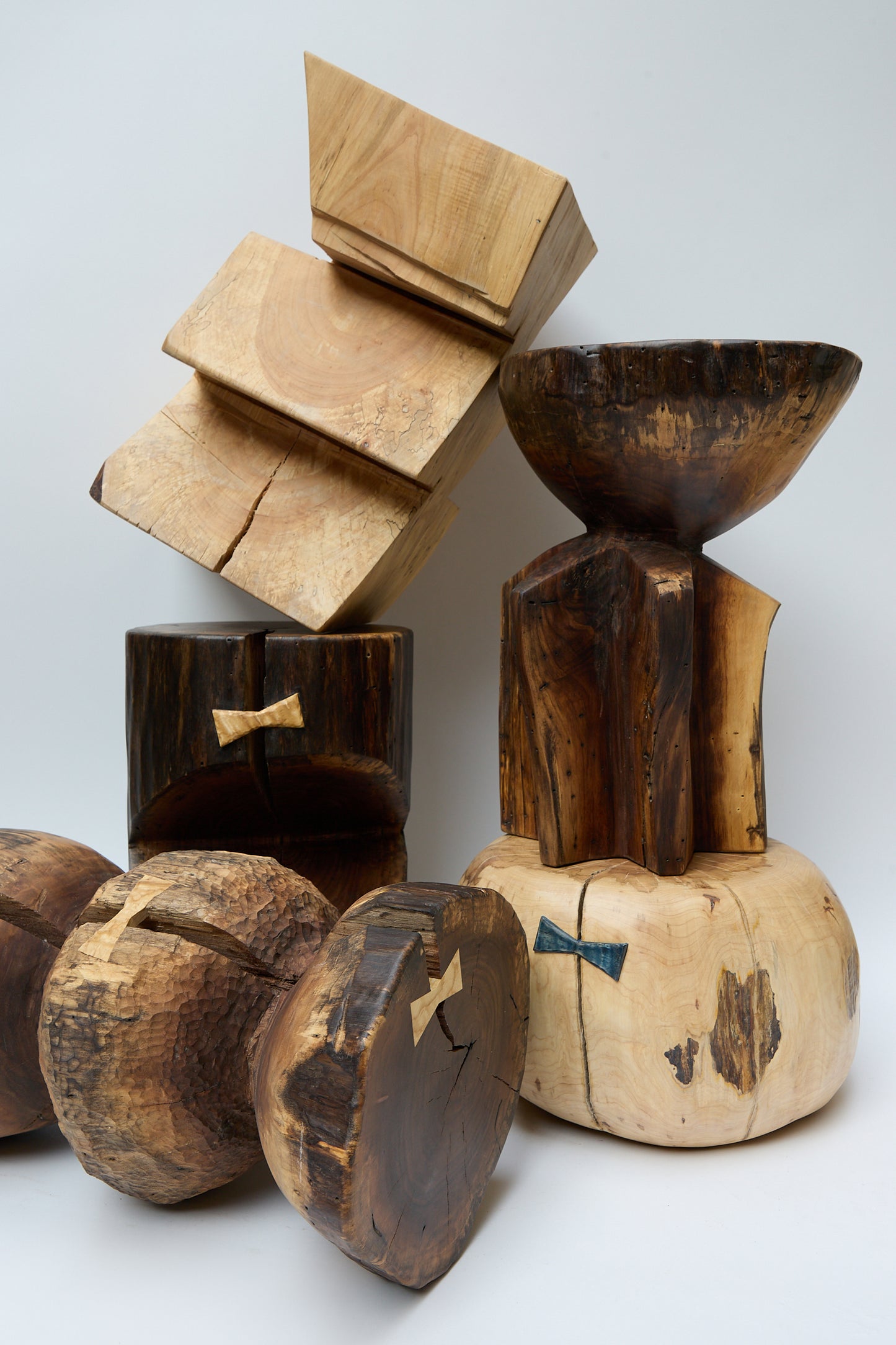 A collection of wooden sculptures with various shapes and sizes, including cube, bowl, and cylindrical forms, displayed against a plain white background. Among them is a stunning **Kingston Walnut VI Side Table** crafted from the rich wood of a walnut tree by **Cody Brgant**.