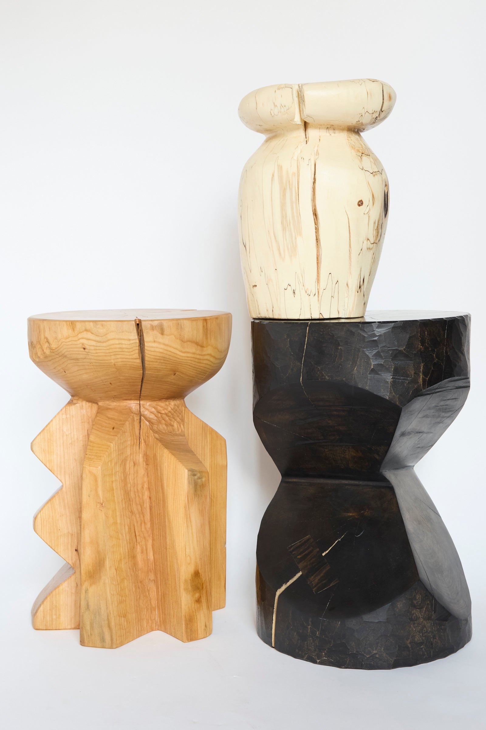 Three wooden sculptural side tables, feature different shapes and colors: one is the Starburst Side Table in Hogback Cherry by Cody Brgant with its light brown and jagged edges, another is smooth black, and a third has a cream color with rounded features. 