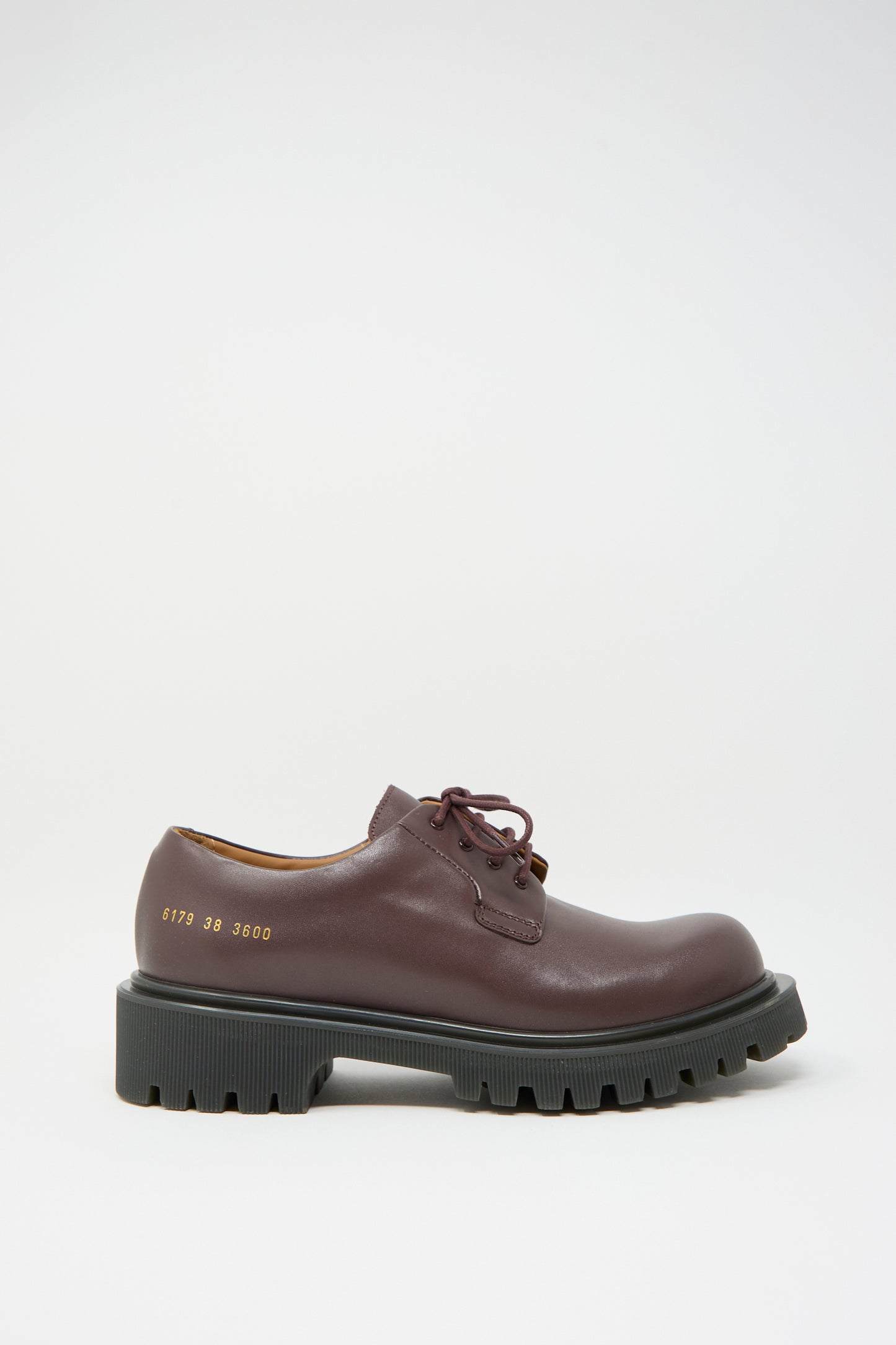 The Chunky Derby Article 6179 in Burgundy with Gum by Common Projects is a luxurious burgundy leather Derby shoe, made in Italy, featuring thick black lug soles and laces, adorned with gold-embossed numbers on the side.