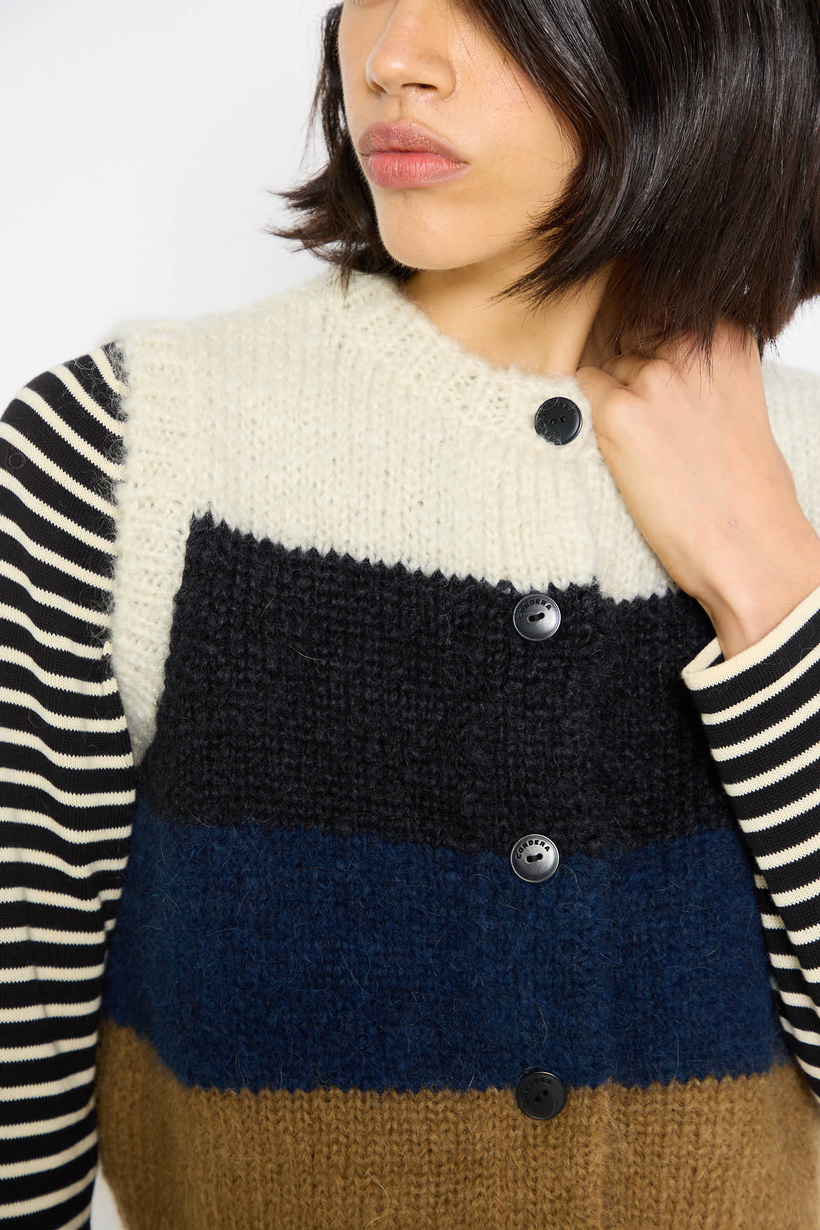 A person with short black hair is wearing Cordera's Alpaca and Mohair Waistcoat in Striped, featuring horizontal stripes in black, white, blue, and brown with black buttons, over a black and white striped shirt.