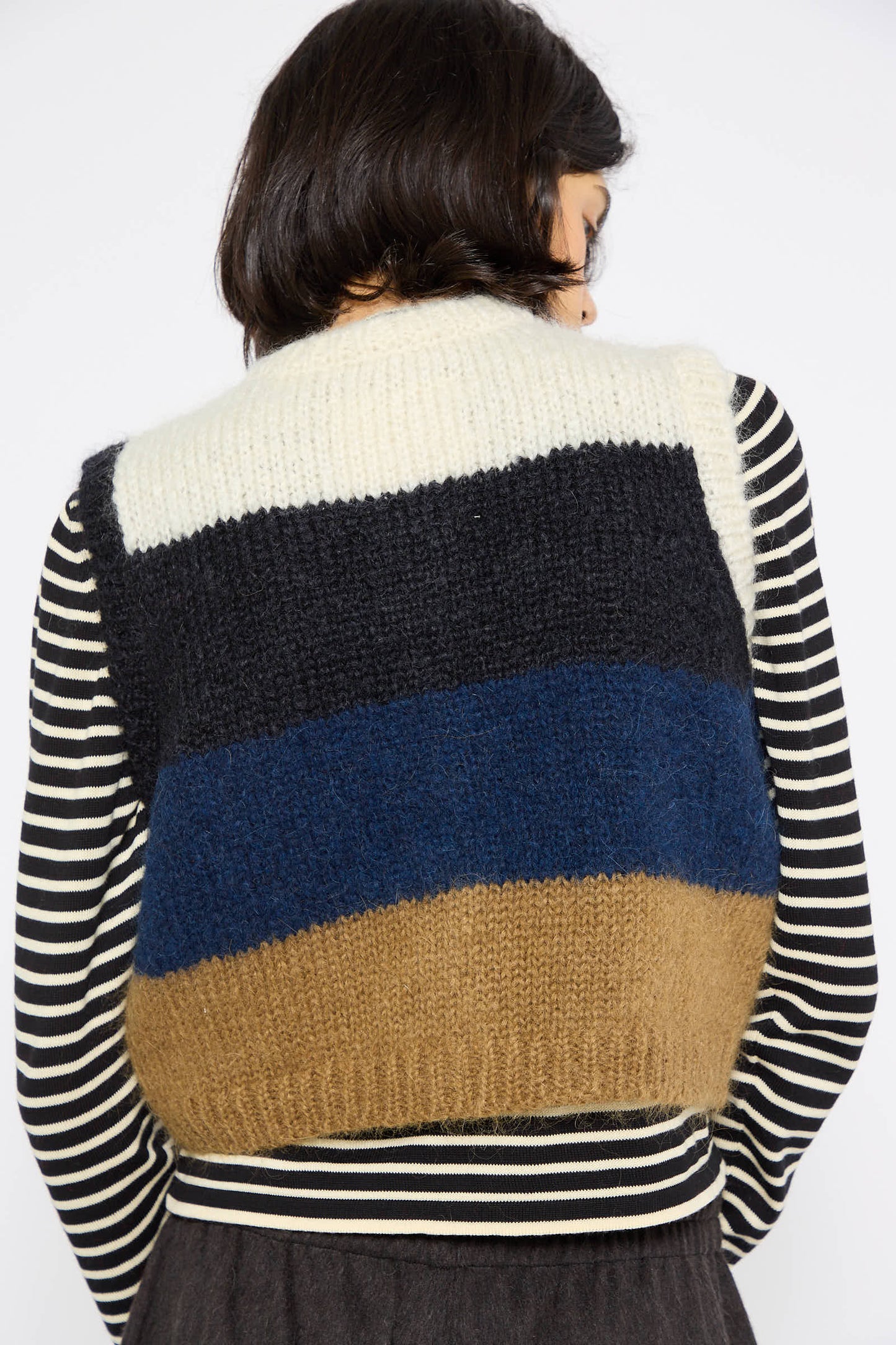 A person with short dark hair wears Cordera's Alpaca and Mohair Waistcoat in Striped over a black and white striped shirt. The ethically manufactured waistcoat, made from a mohair blend, features horizontal stripes in white, black, blue, and brown. The person is facing away.