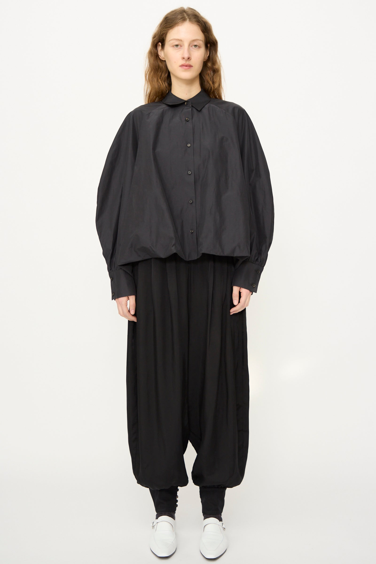 A person faces forward in a Cordera Balloon Shirt in Black with voluminous sleeves, wide black pants, black leggings, and white shoes, all set against a plain white background.