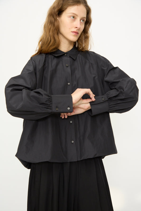 A person with long brown hair wears Cordera's Balloon Shirt in Black with voluminous sleeves and black pants, standing against a plain white background.