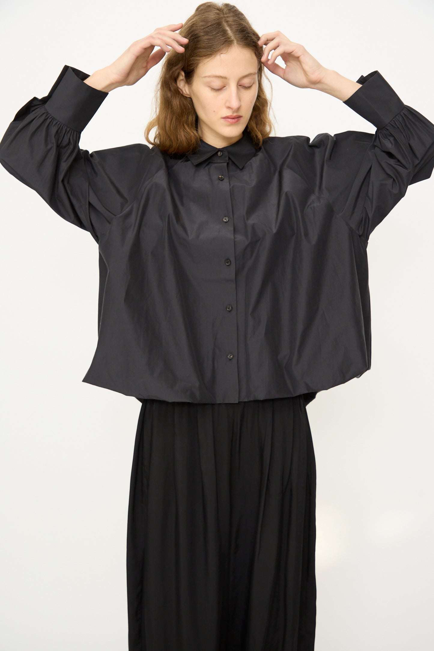 A person with long hair wears the Cordera Balloon Shirt in Black and black pants, standing against a plain white background with eyes closed and hands raised near their head.