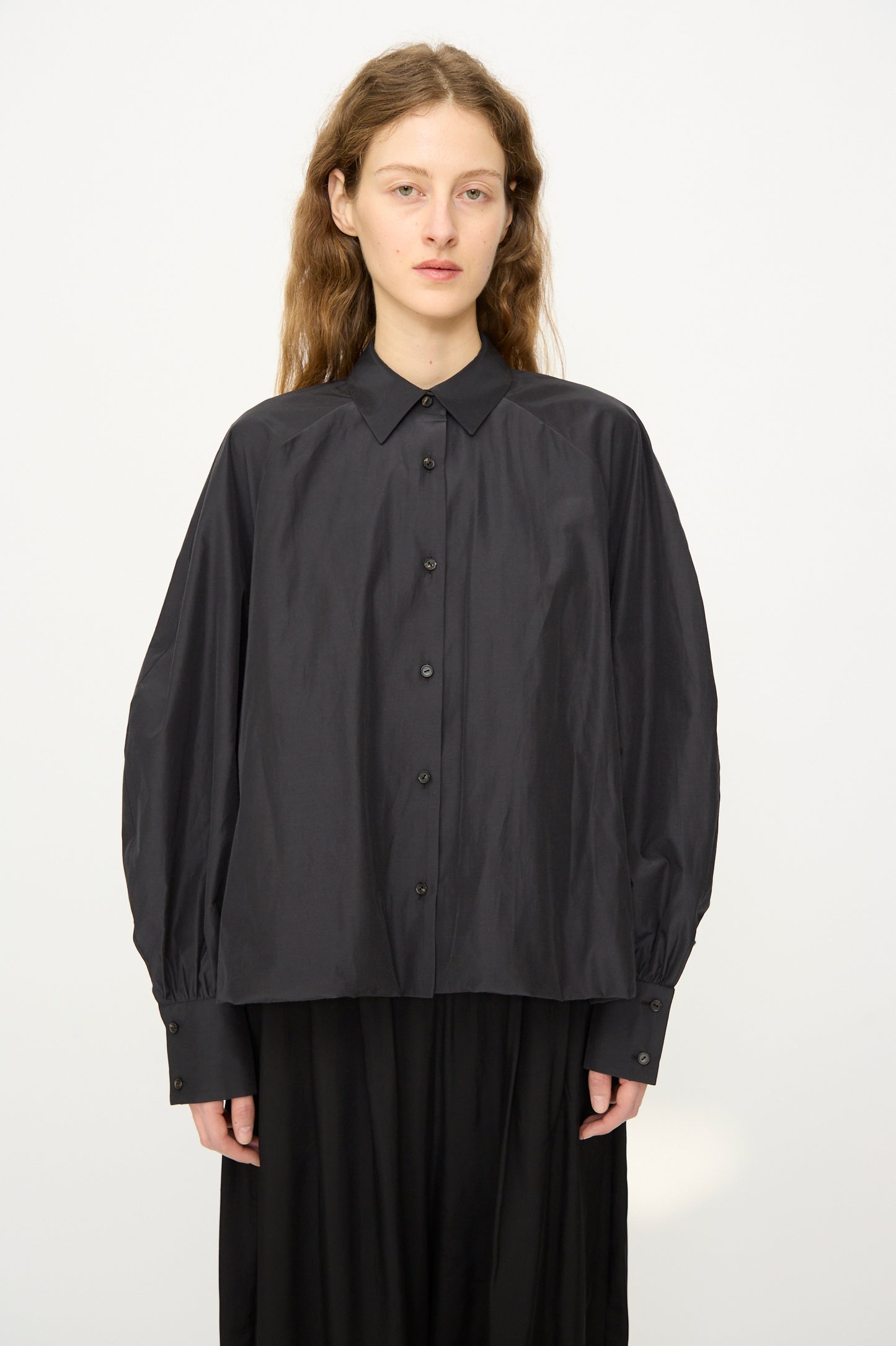 A person with long hair wears the Cordera Balloon Shirt in Black, featuring sheer fabric and voluminous sleeves, paired with a black skirt, standing against a plain white background.
