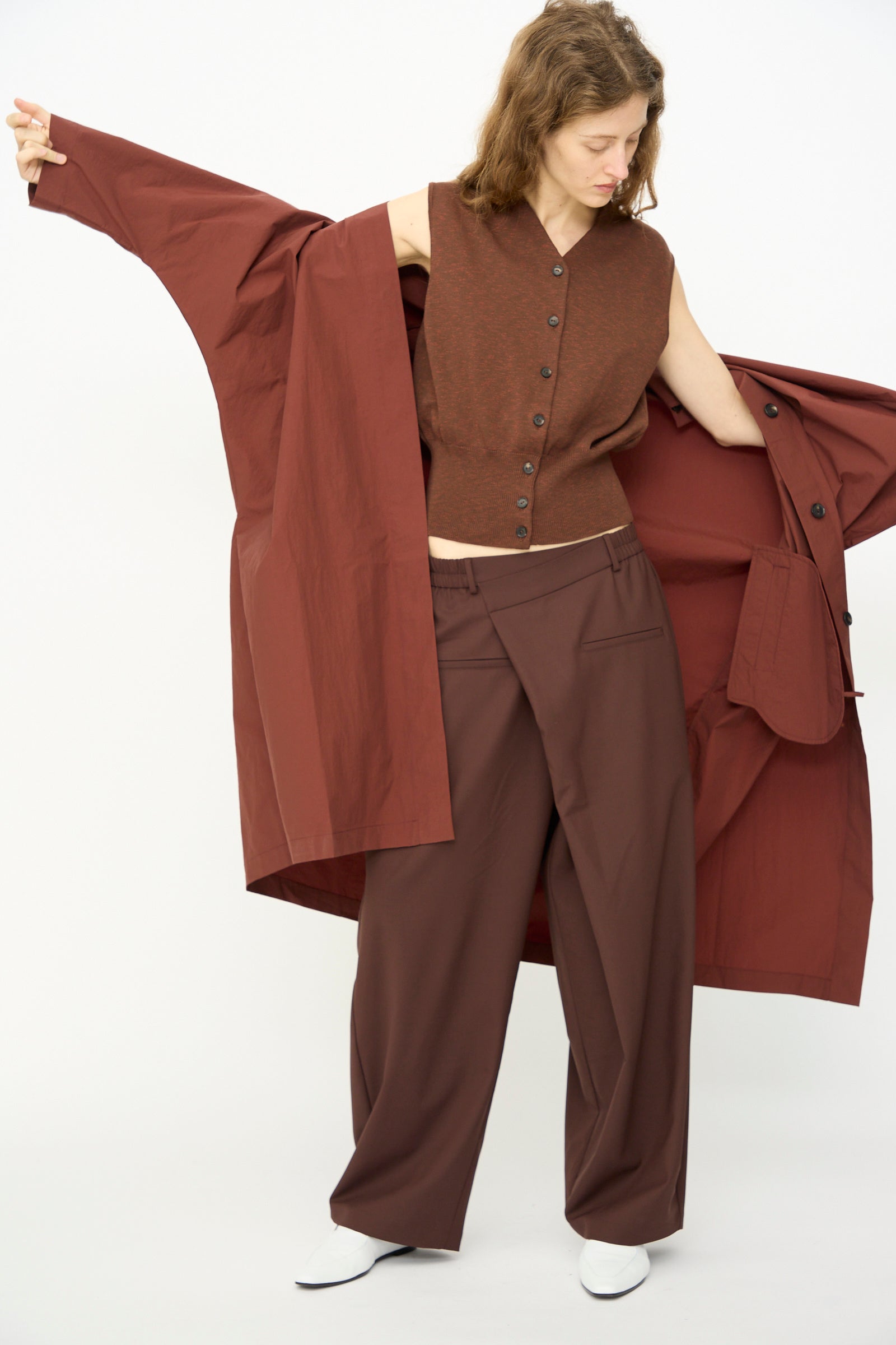 A person stands against a white background wearing Cordera's Batwing Trench in Sienna—an oversized, open long coat—paired with wide-leg brown trousers, a sleeveless brown vest, and white shoes.