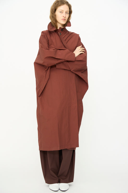 A person stands with arms crossed, wearing Cordera's Batwing Trench in Sienna and matching wide-leg pants, paired with white shoes, against a plain white background.