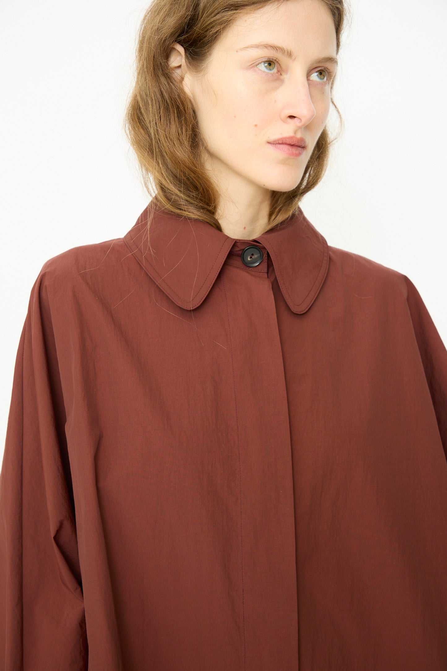 A person with light brown hair wears the Cordera Batwing Trench in Sienna, an oversized brown collared button-up coat, while looking slightly to the side against a plain light background.