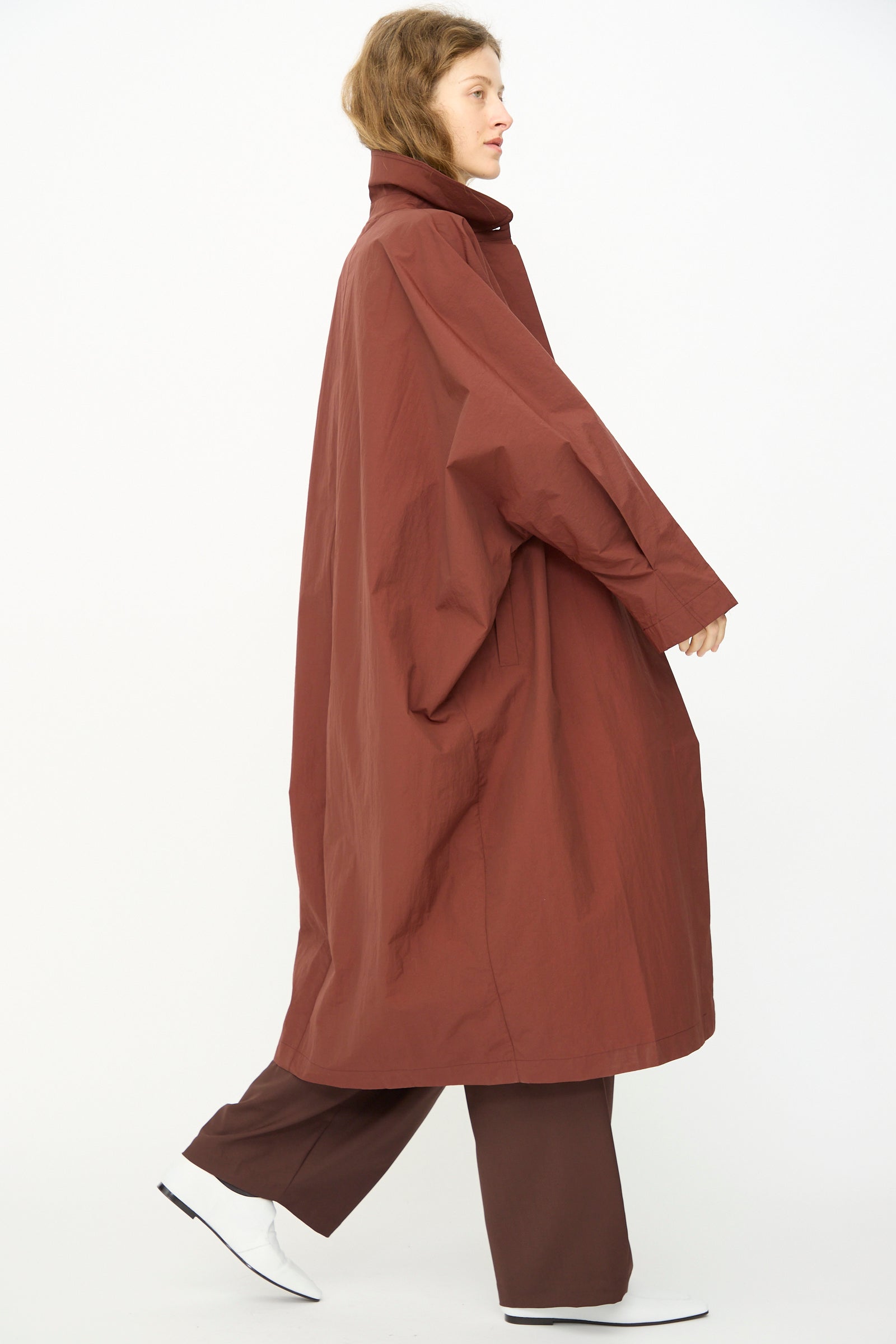 Someone walks against a plain white background wearing Cordera's Batwing Trench in Sienna, paired with brown pants and white shoes.