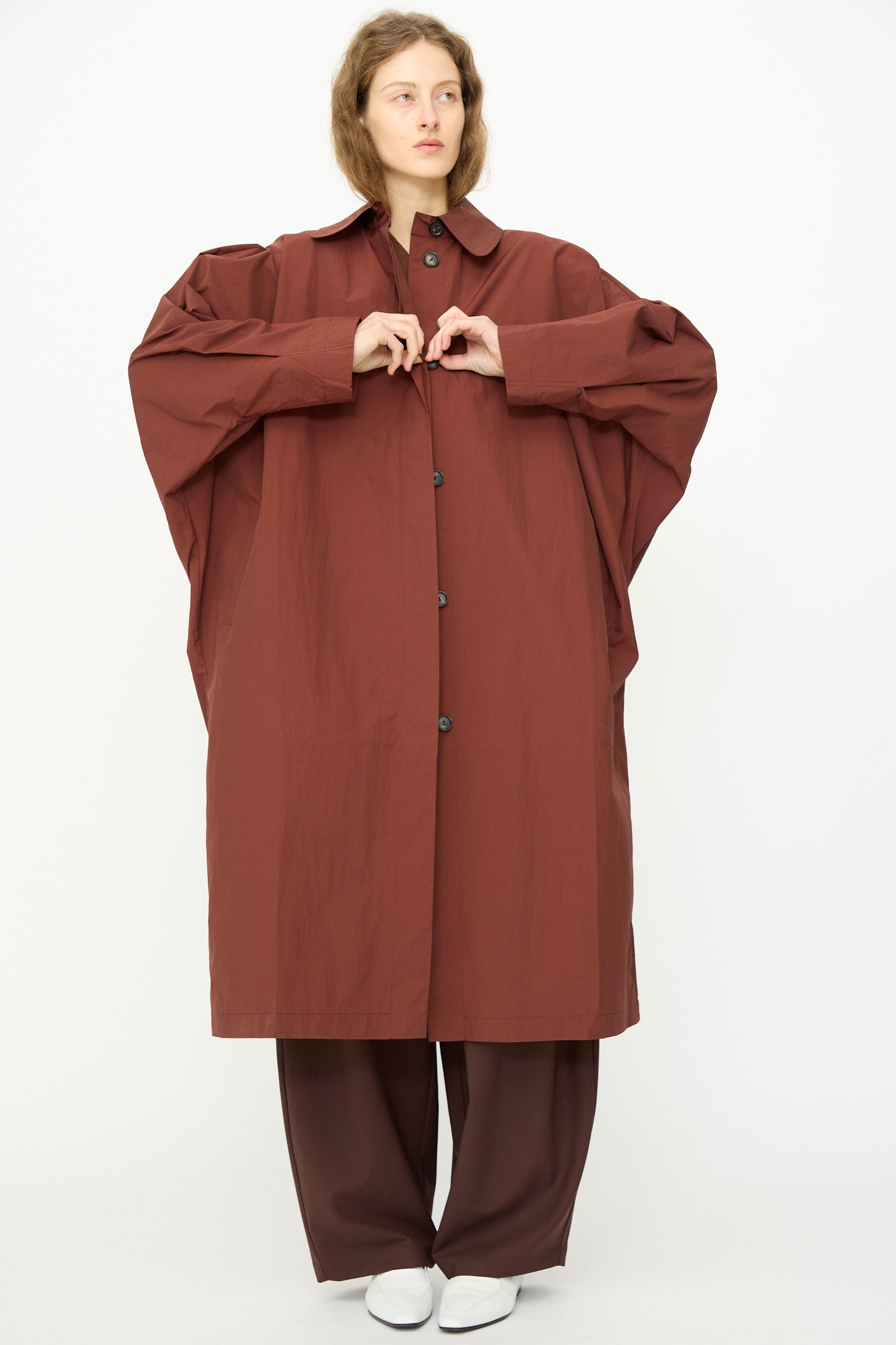 A person stands against a plain background wearing Cordera's Batwing Trench in Sienna, paired with matching wide-leg pants and white shoes.