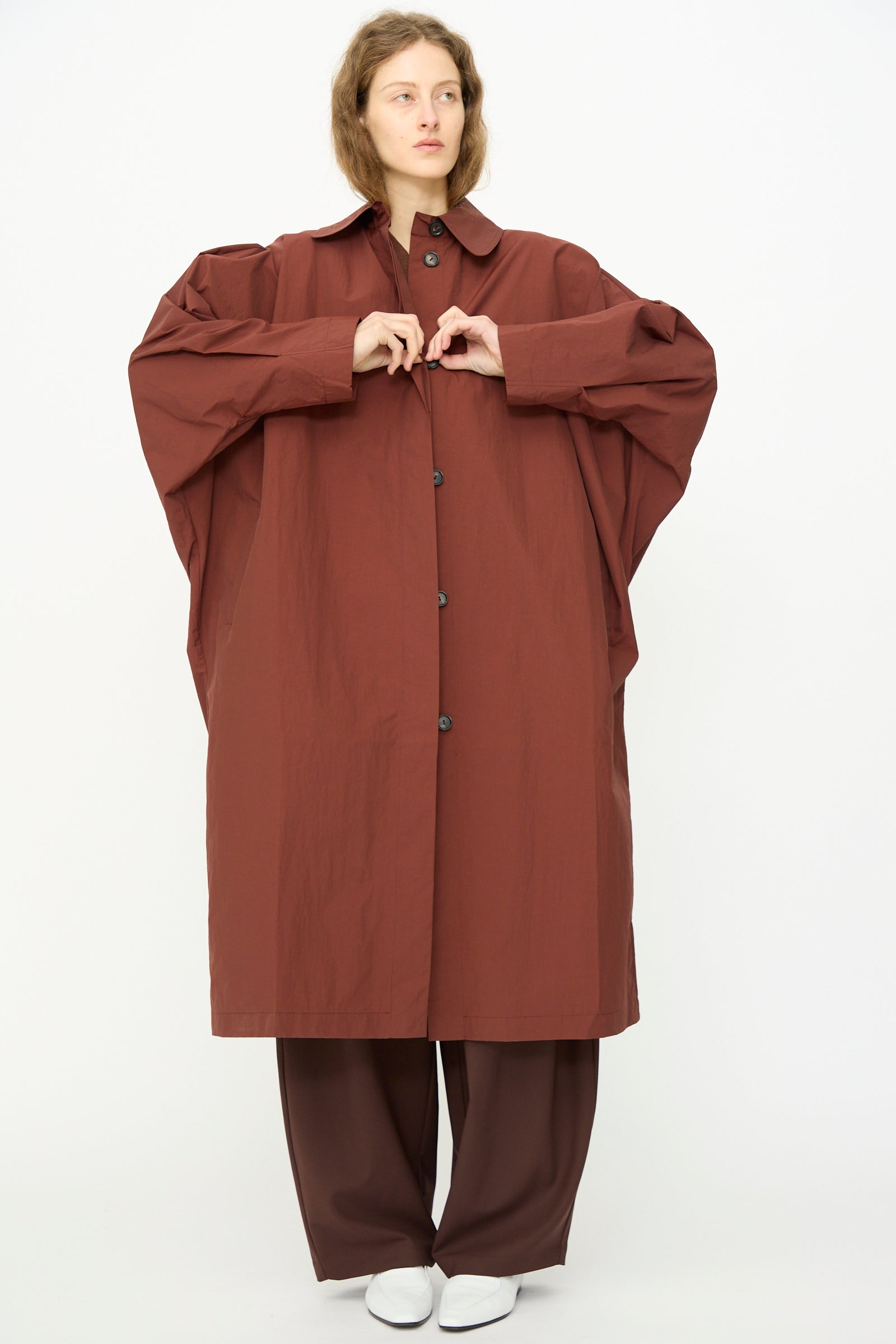 A person stands against a plain background wearing Cordera's Batwing Trench in Sienna, paired with matching wide-leg pants and white shoes.