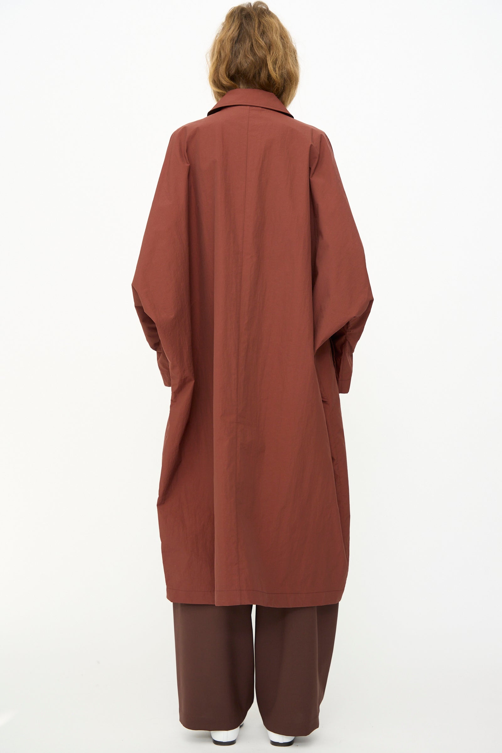 A person with shoulder-length hair wears the Cordera Batwing Trench in Sienna, styled with brown pants, standing with their back to the camera against a plain white background.