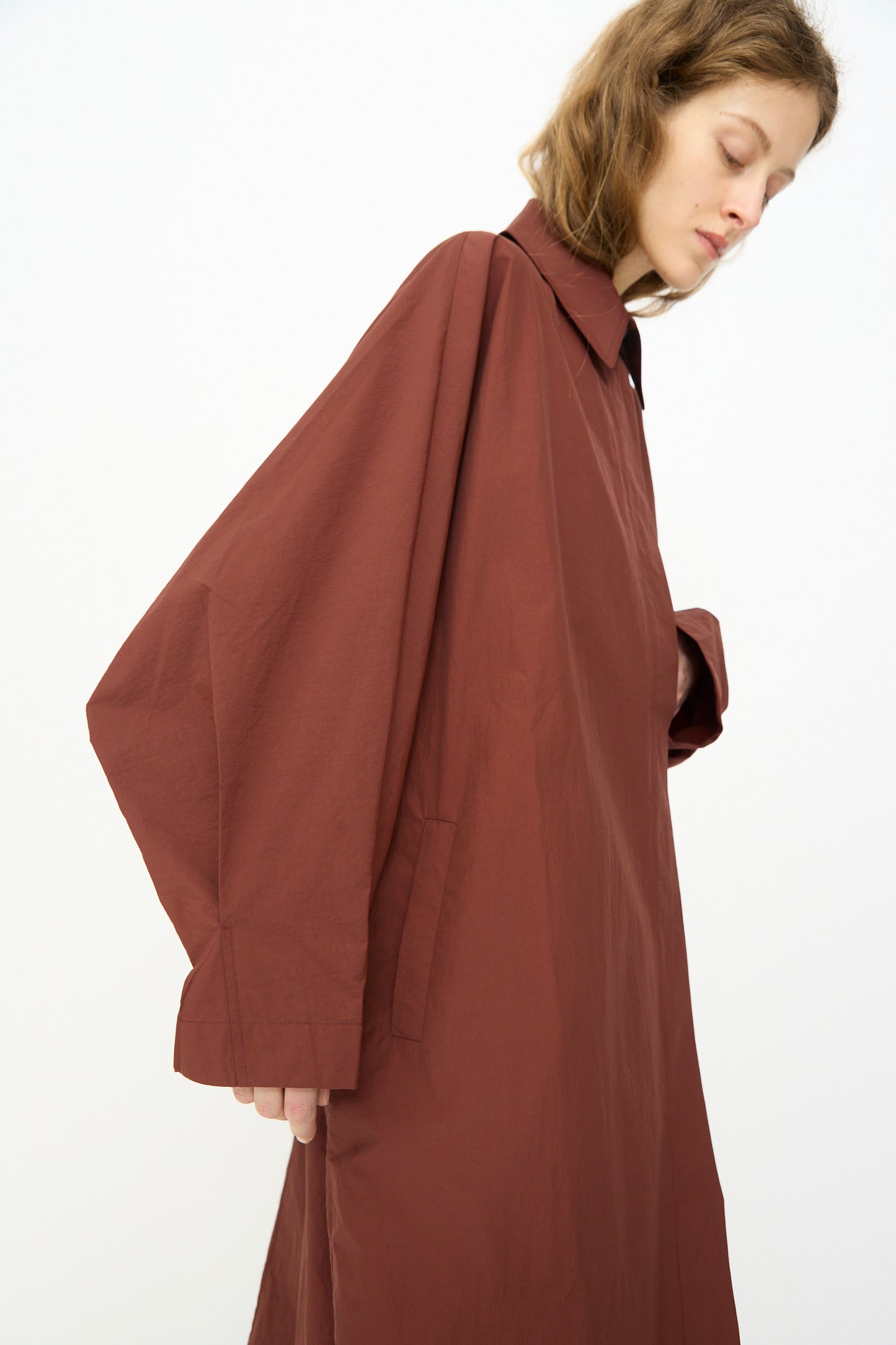 A person wearing the Cordera Batwing Trench in Sienna stands sideways against a plain white background, their head slightly bowed.