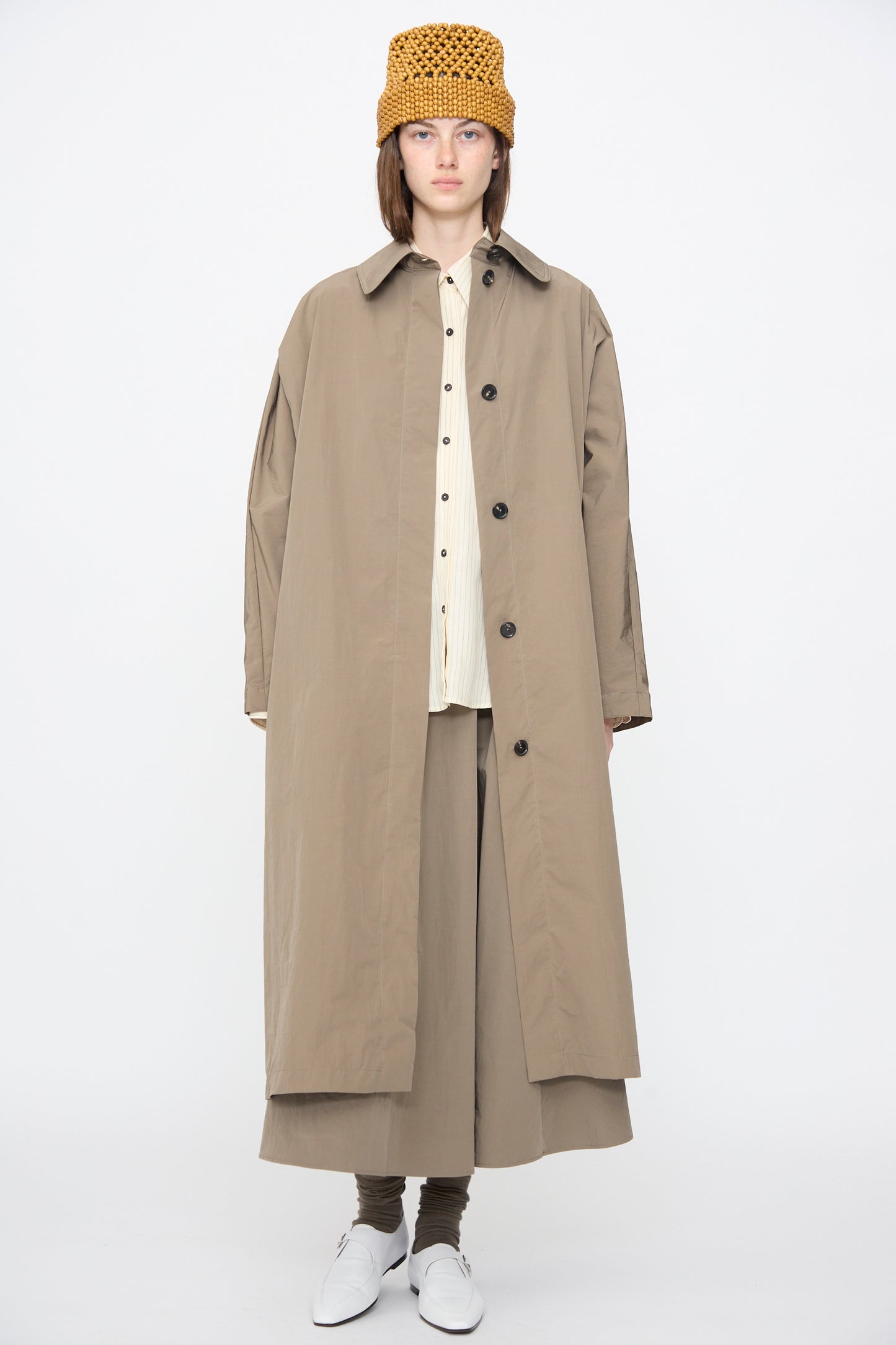 A person stands facing forward in Cordera's Batwing Trench in Vetiver, paired with tan wide-leg pants, a white shirt, mustard crochet hat, white shoes, and brown socks against a plain white background.