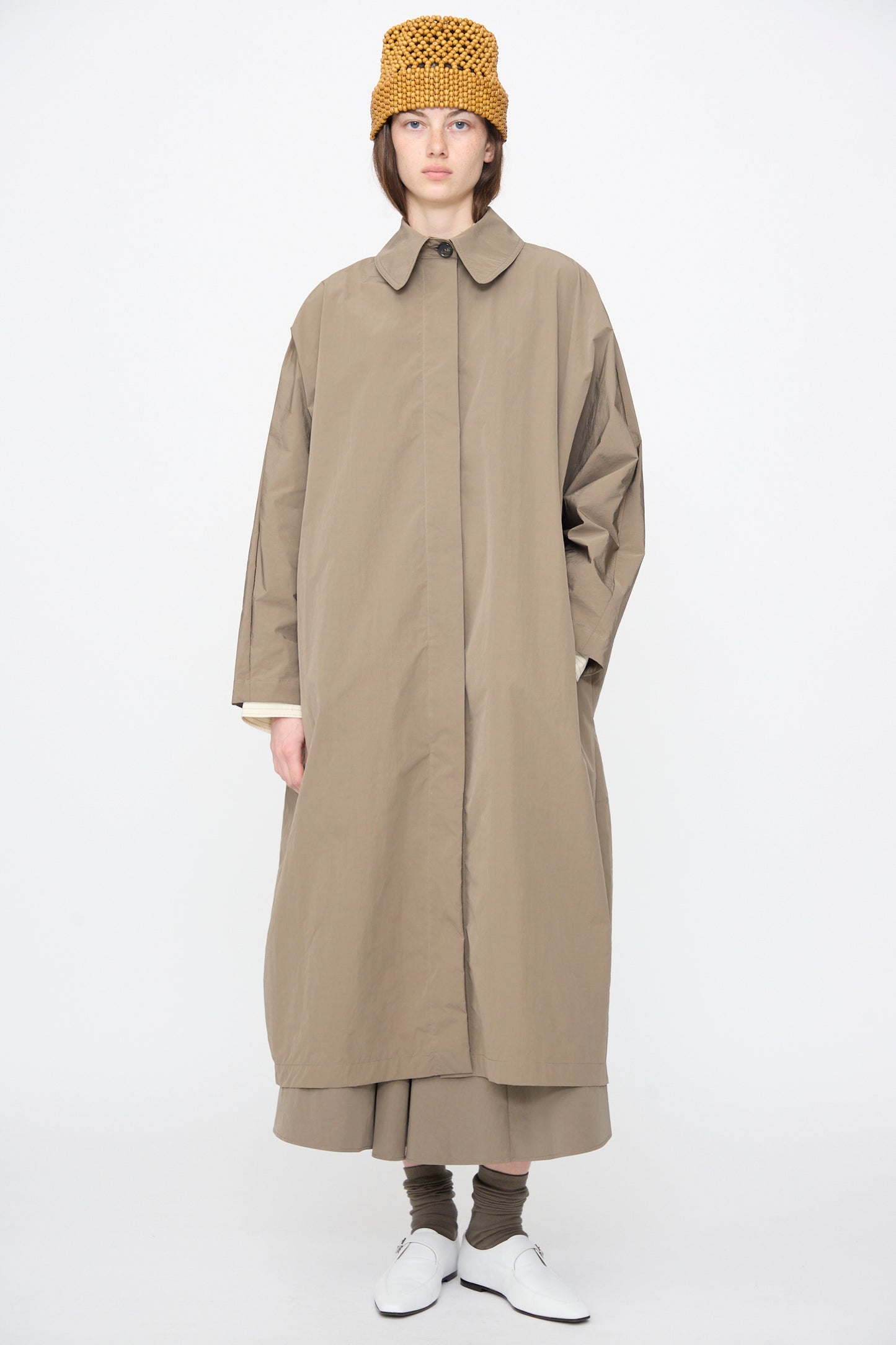 A person wearing Cordera’s Batwing Trench in Vetiver with a matching skirt, brown socks, white shoes, and a yellow crochet hat stands against a plain white background.