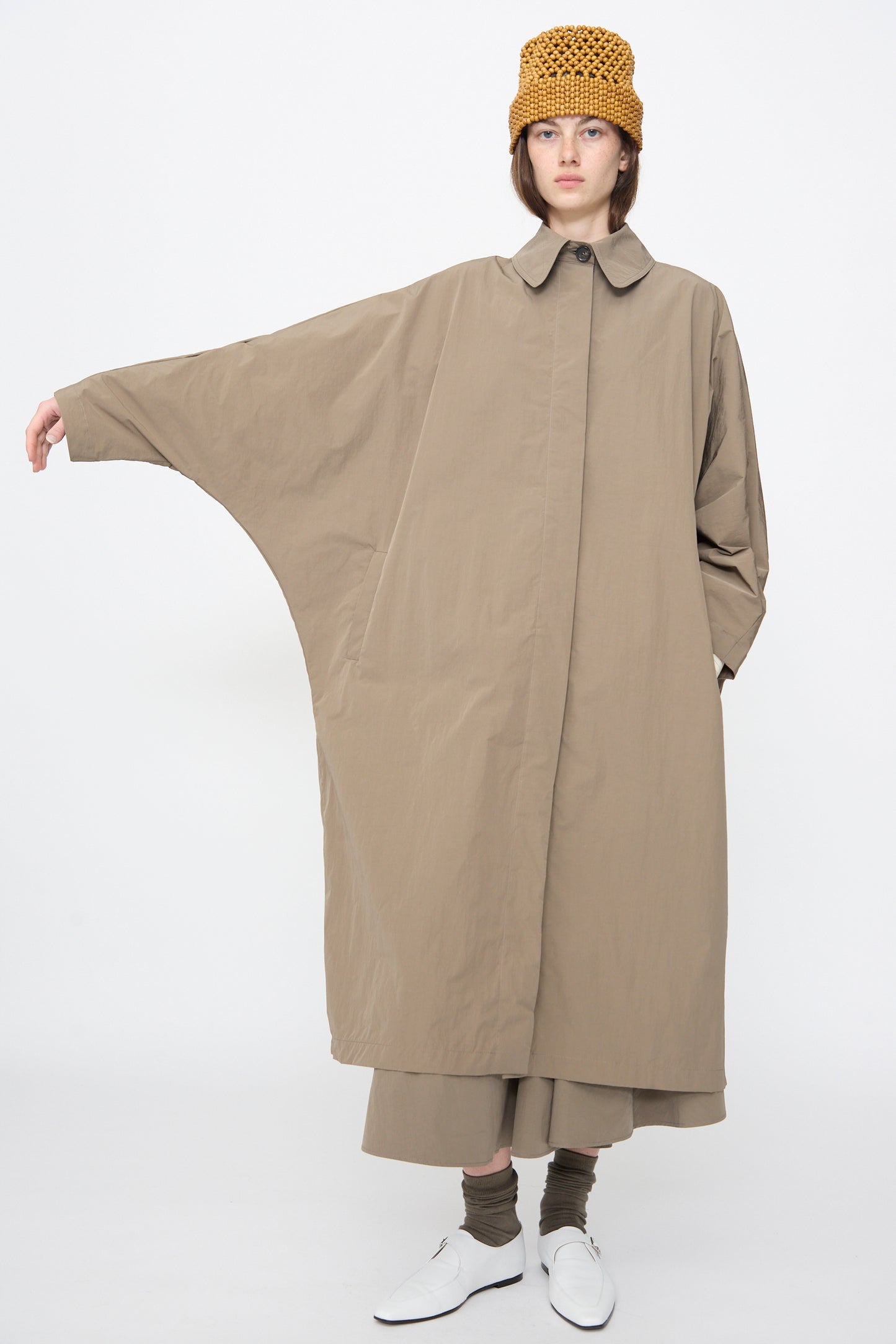 A person wears Cordera's Batwing Trench in Vetiver—a cotton blend coat with wide batwing sleeves—paired with a mustard knit hat, wide pants, white shoes, and gray socks, standing against a plain white background.
